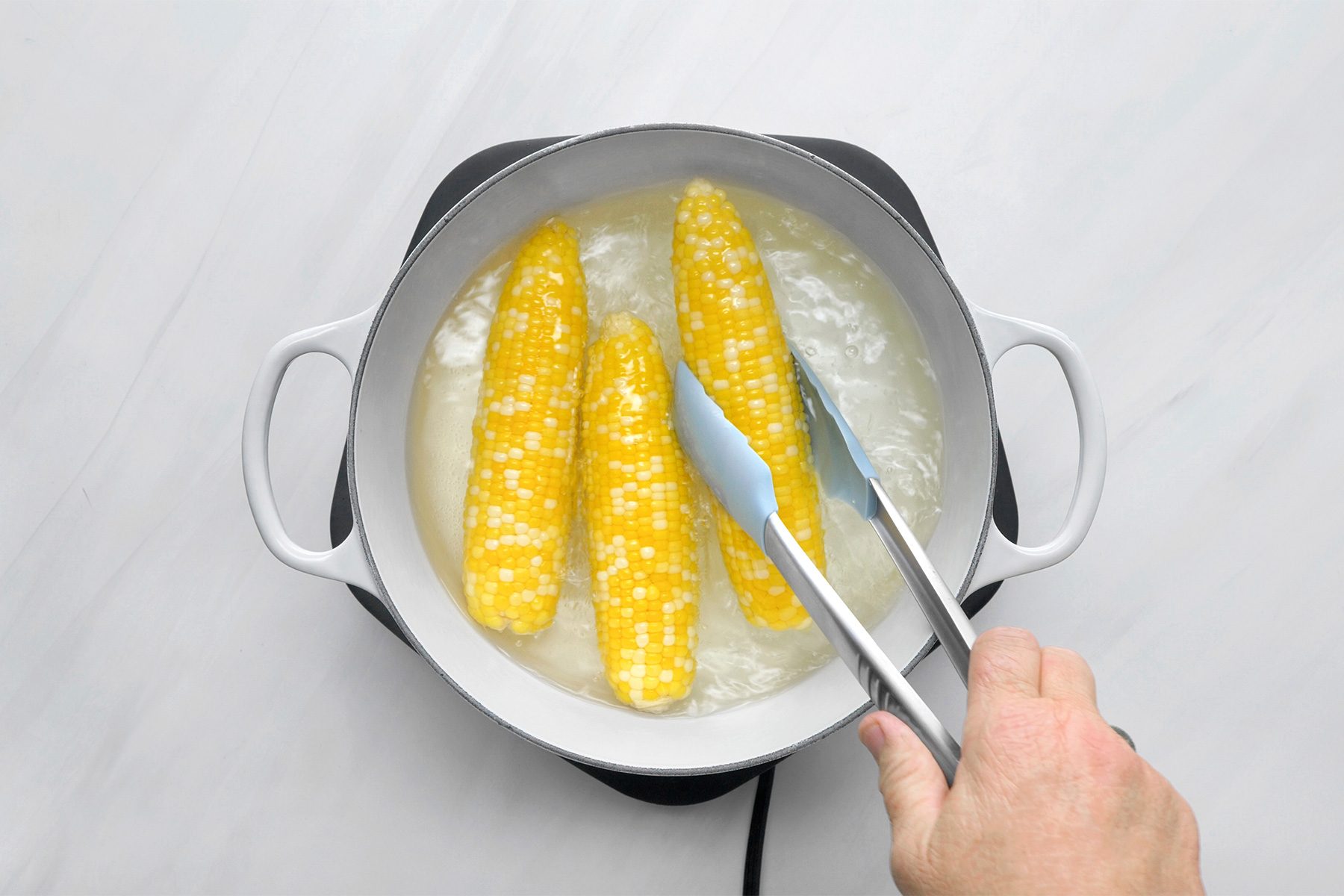 overhead shot of a white pot filled with boiling water, Several ears of corn are submerged in the water, with their husks removed, A pair of blue tongs is being used to turn one of the corn cobs, ensuring even cooking;