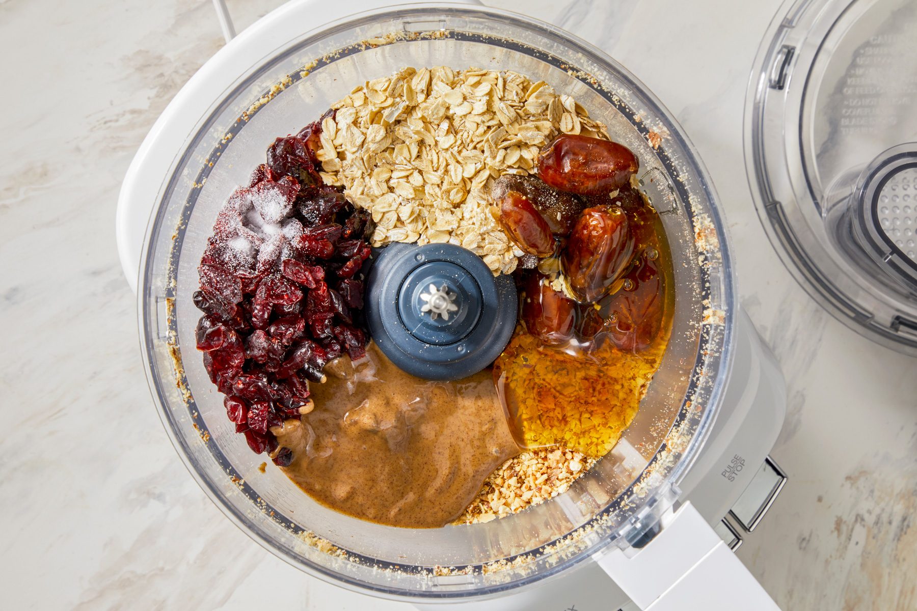 A food processor bowl filled with finely chopped almonds, with dates, dried cranberries, rolled oats, almond butter, honey, salt, and almond extract added on top. The ingredients are layered, ready to be blended together.
