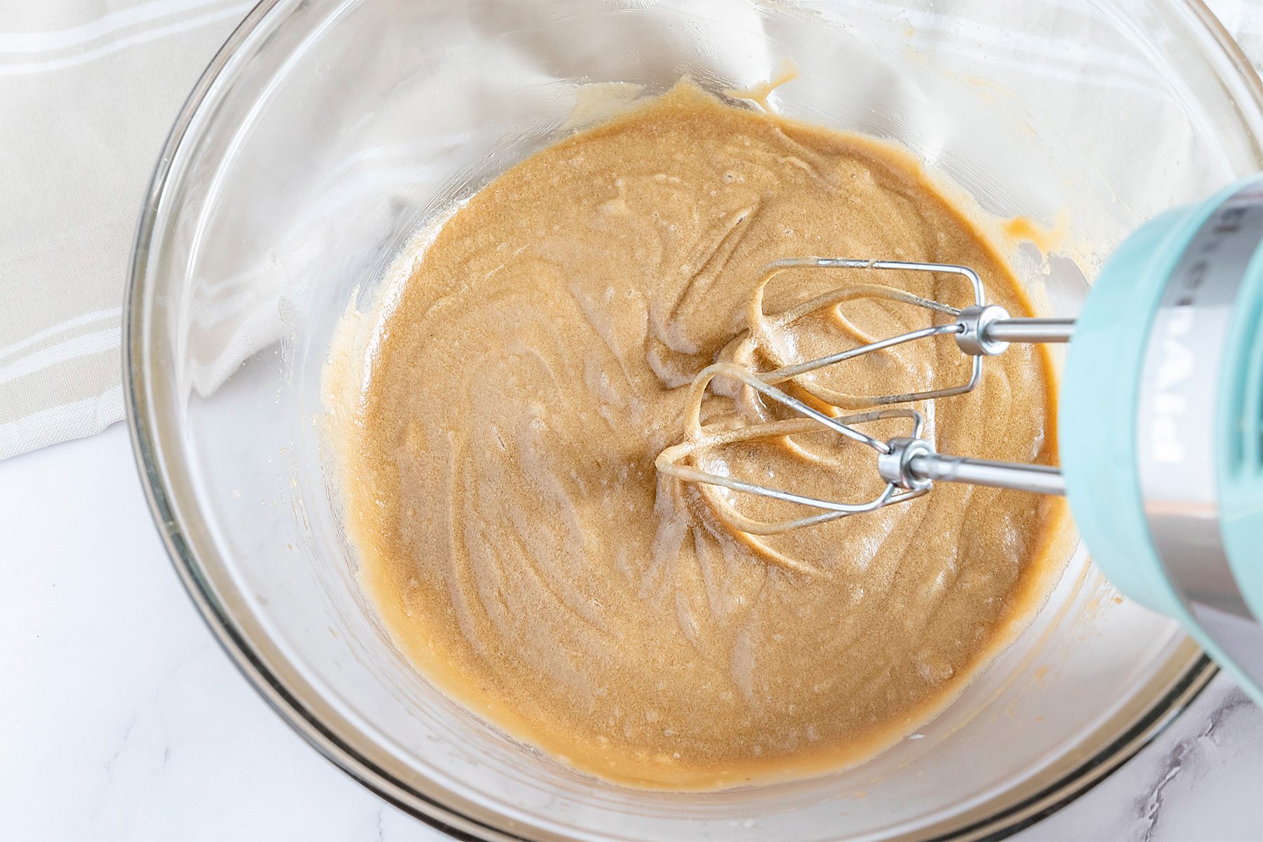 A mixing bowl with creamy peanut butter being blended by a hand mixer on a white marble countertop.