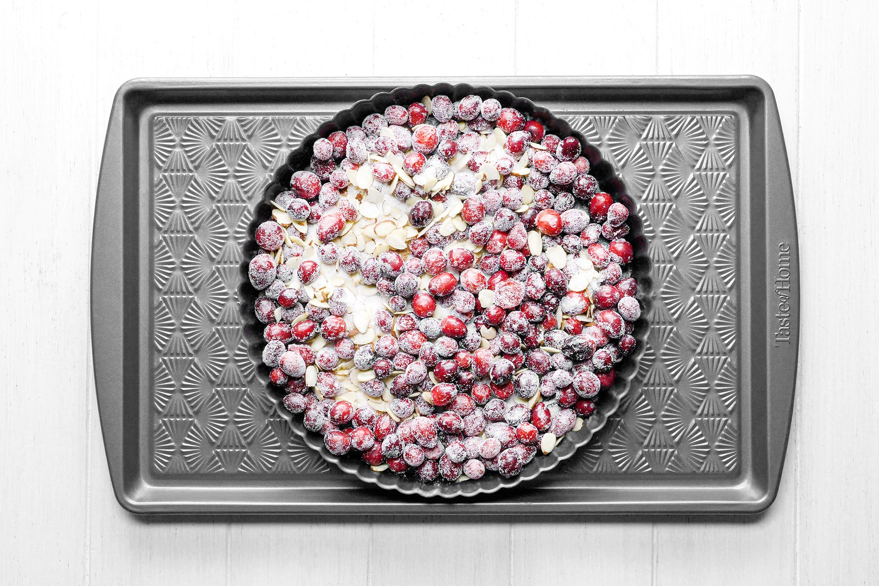 A round tart pan filled with unbaked cranberry pie with sugar and almond slices is placed on a rectangular baking sheet with a textured pattern, set on a white surface.