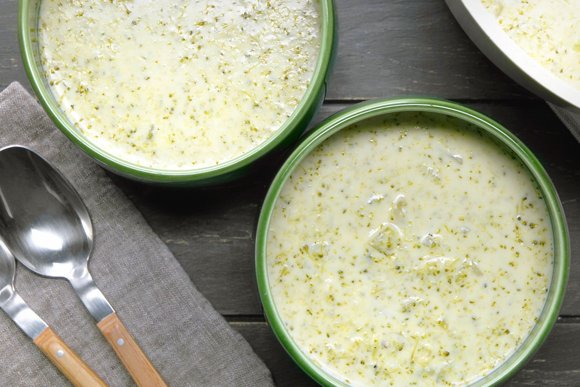 overhead shot of Two green bowls filled with Creamy Broccoli Soup are showcased, The soup is a creamy, pale yellow color with visible flecks of broccoli, The texture appears thick and smooth, Spoons are placed near the bowls;