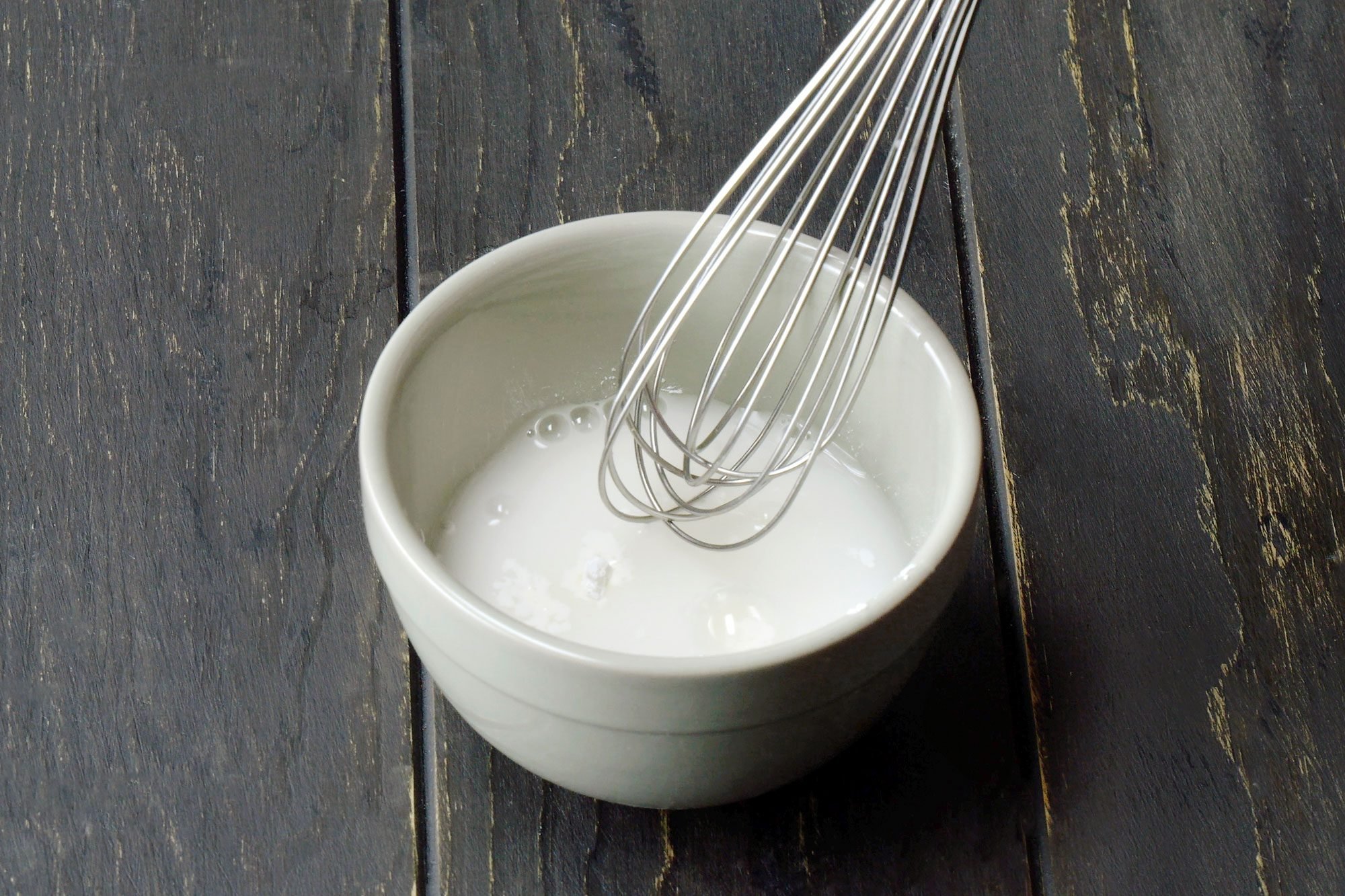 3/4th shot of a white bowl contains a liquid mixture that is being whisked with a metal whisk, The liquid appears to be a light yellow color and has bubbles forming on the surface, The bowl is placed on a black wooden surface;