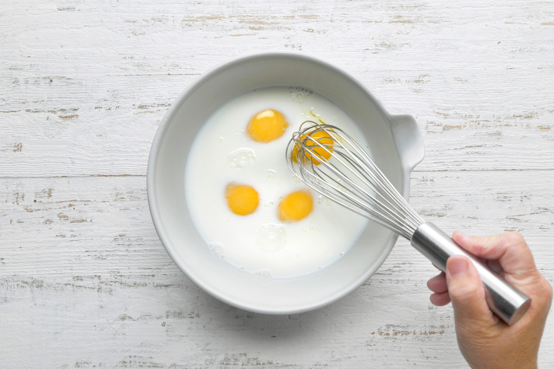 a top-down view of a white bowl on a white wooden surface, Inside the bowl, there are three whole eggs with intact yolks surrounded by clear egg whites, A hand is holding a whisk, partially submerged in the egg whites