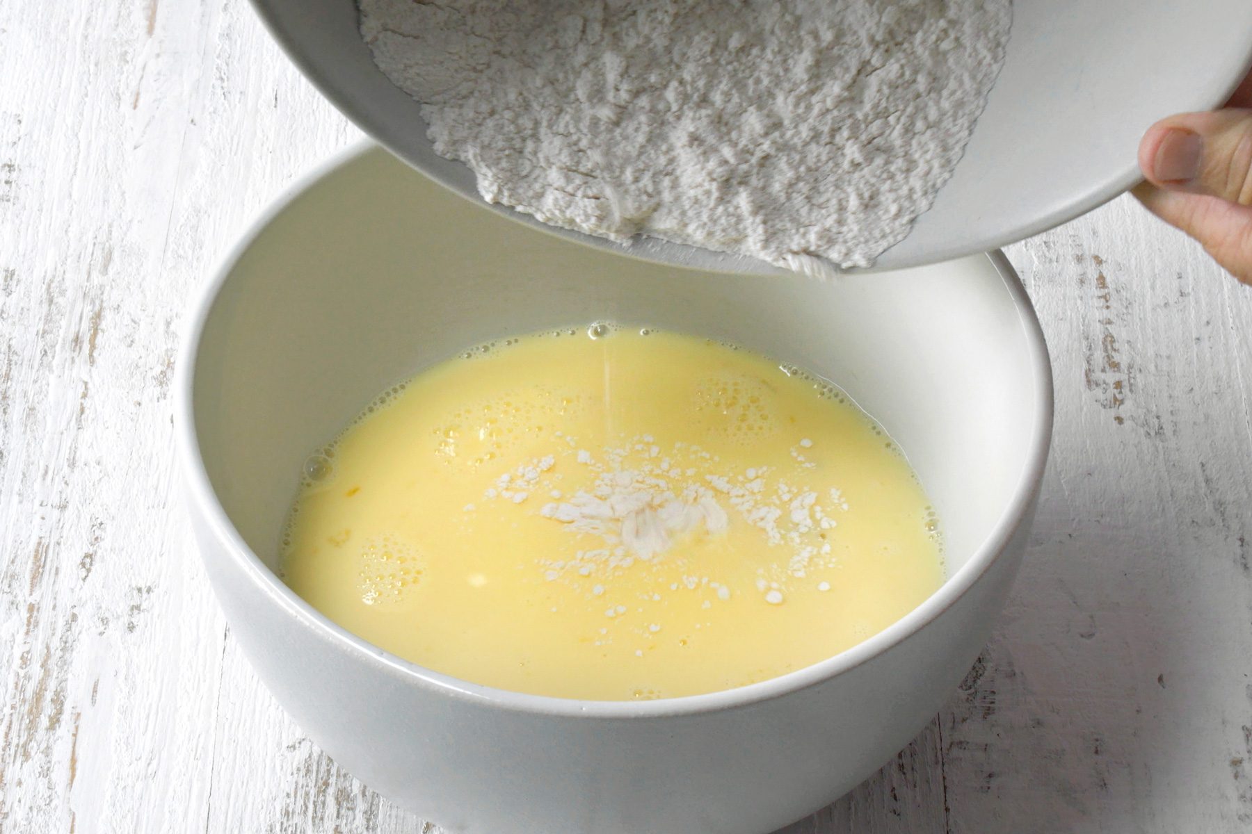 a person’s hand pouring a white powdery substance, likely flour, into a white bowl containing a yellow liquid mixture, which appears to be beaten eggs or batter