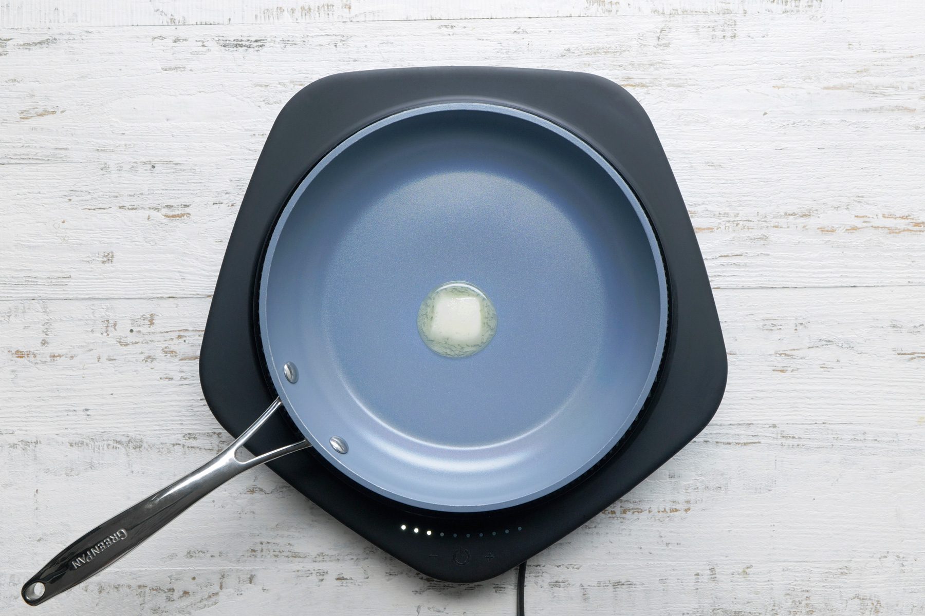 overhead shot; white wooden background; a blue frying pan with a silver handle, placed on a black square plate, Inside the pan, there’s a small pat of butter melting in the center