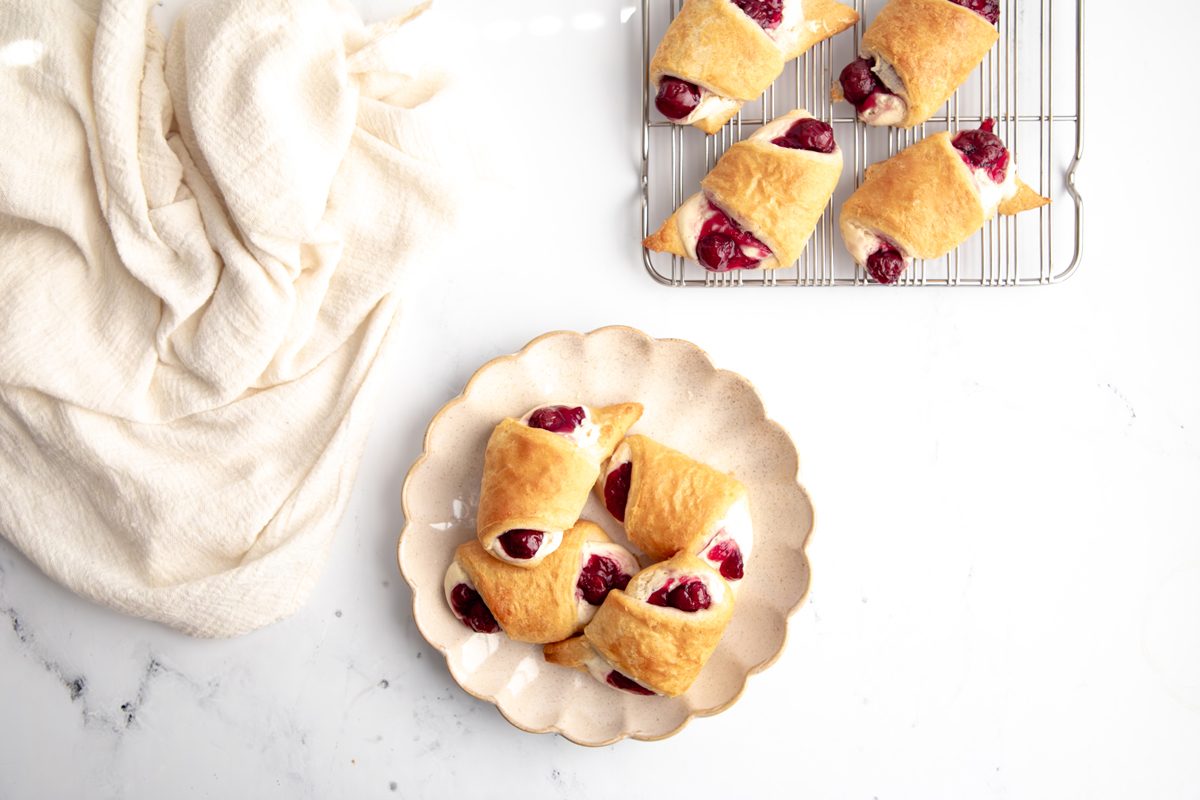 Overhead Beauty shot for Taste of Crescent Rolls with Cherry Pie Filling, baked crescent rolls with cream cheese and cherry filling inside on a scalloped plate with additional rolls on a wire rack.