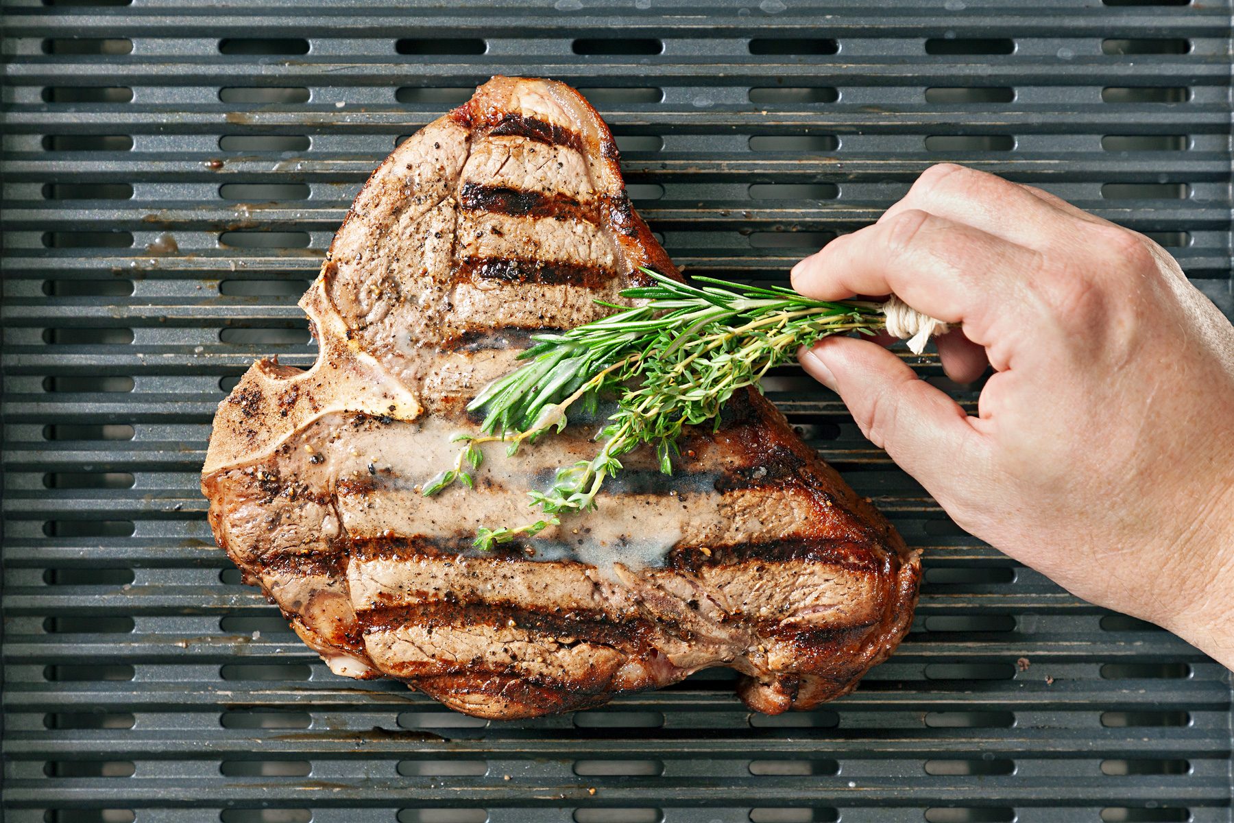 The steak is being basted by dipping a bundle of thyme and rosemary into melted butter and brushing it onto the surface of the meat.
