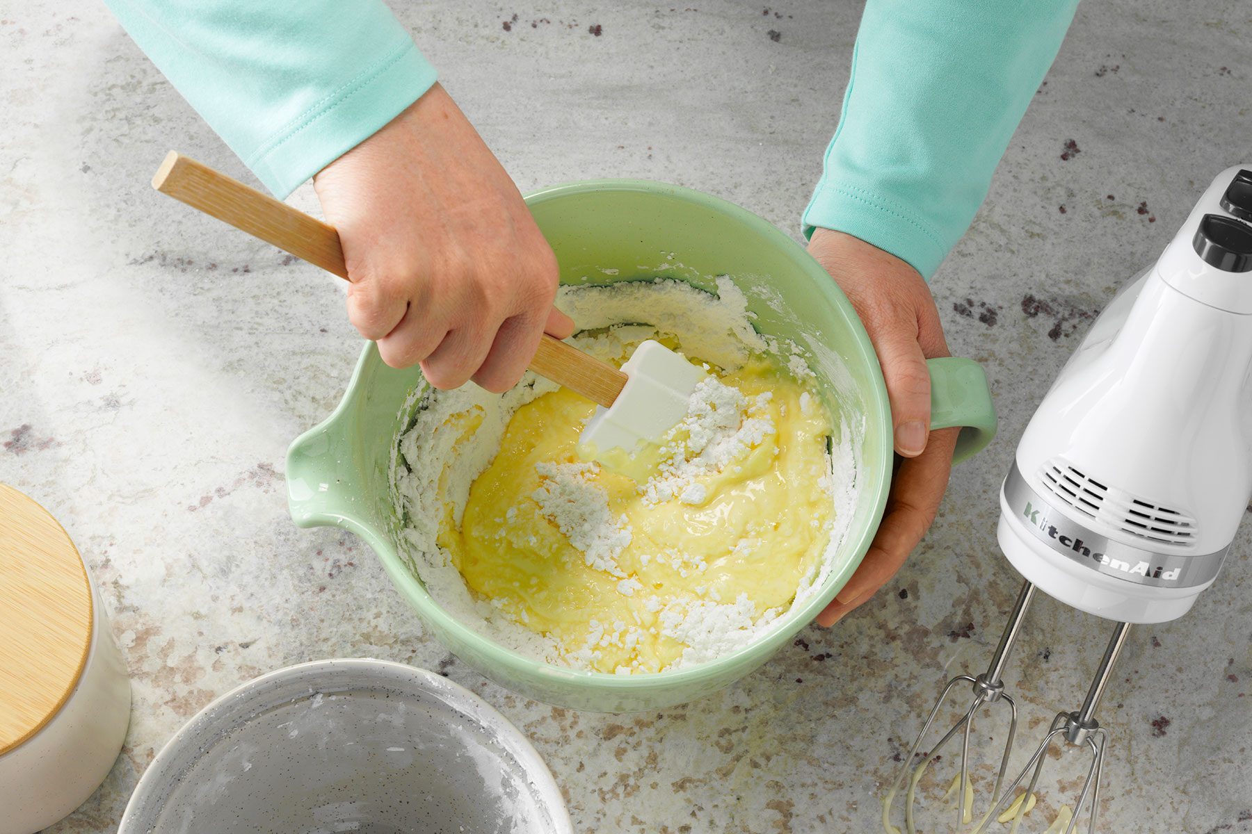 A person in a light blue sweater mixes cake batter in a light green bowl with a spatula. An electric hand mixer and other kitchen items are on the countertop nearby.