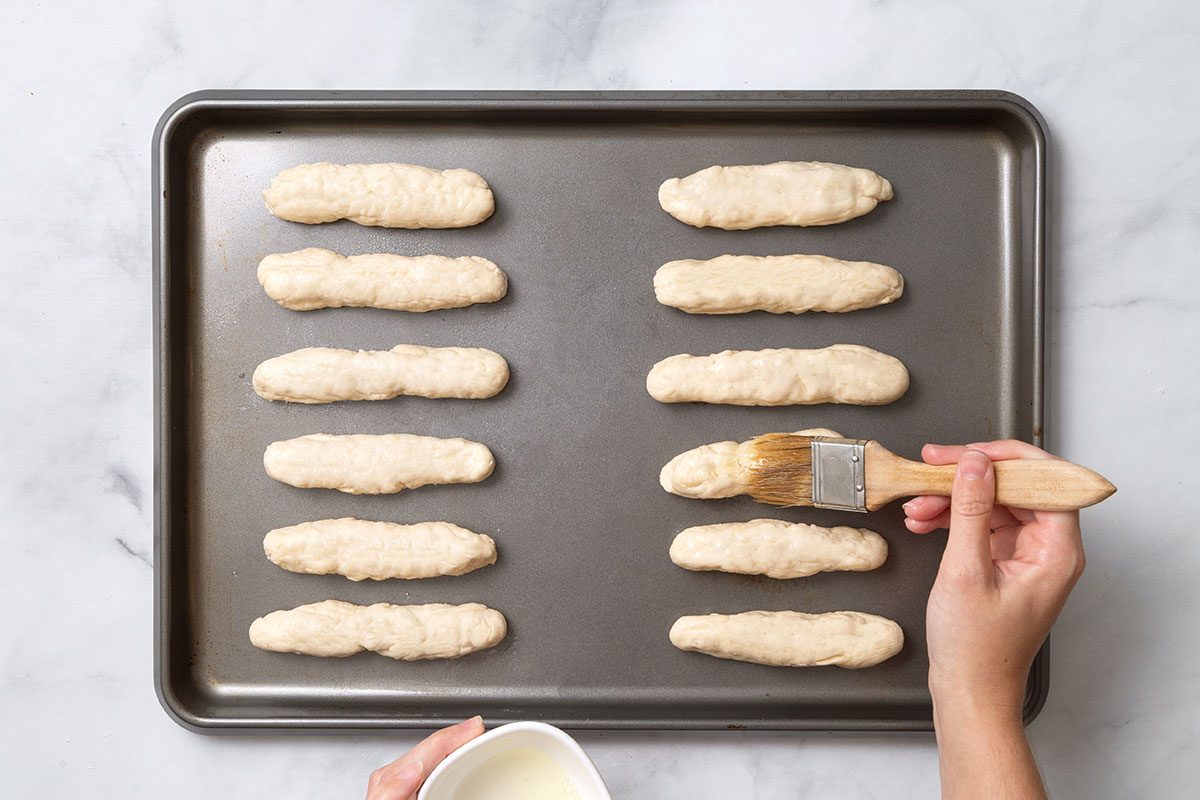 6 inch ropes of bread dough brushed with egg wash on a baking sheet for step five of Homemade Breadsticks for Taste of Home