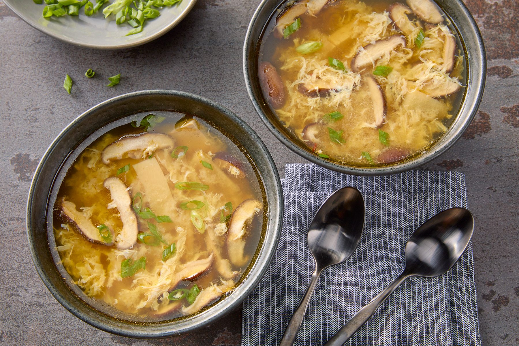 Overhead shot of Easy Hot and Sour Soup; serve soup in bowls; top with green onion; spoons; napkin; brown texture background;