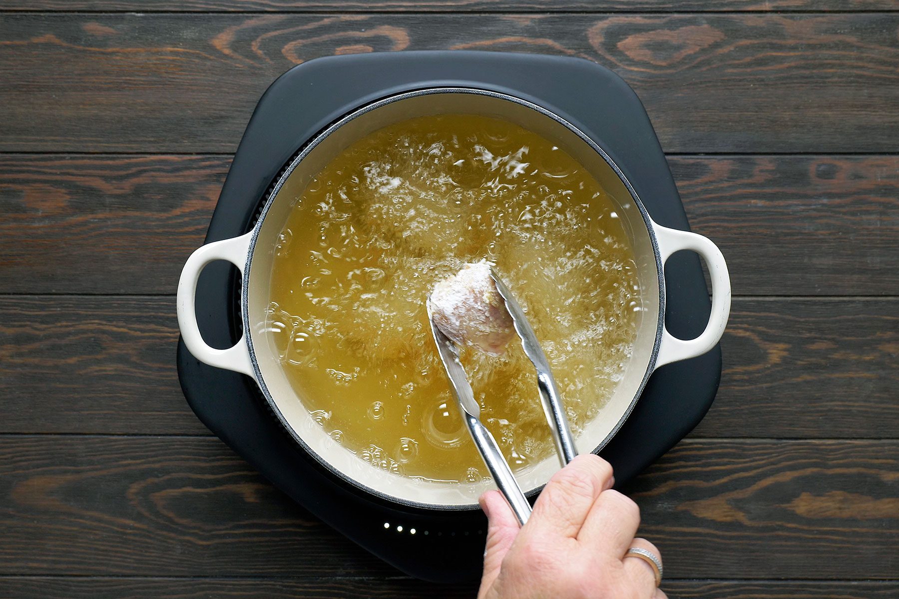 A person uses tongs to fry a coated food item in a pot of hot oil on a stovetop. The oil bubbles vigorously, indicating high heat. The pot is white with side handles, set on a dark wooden surface.