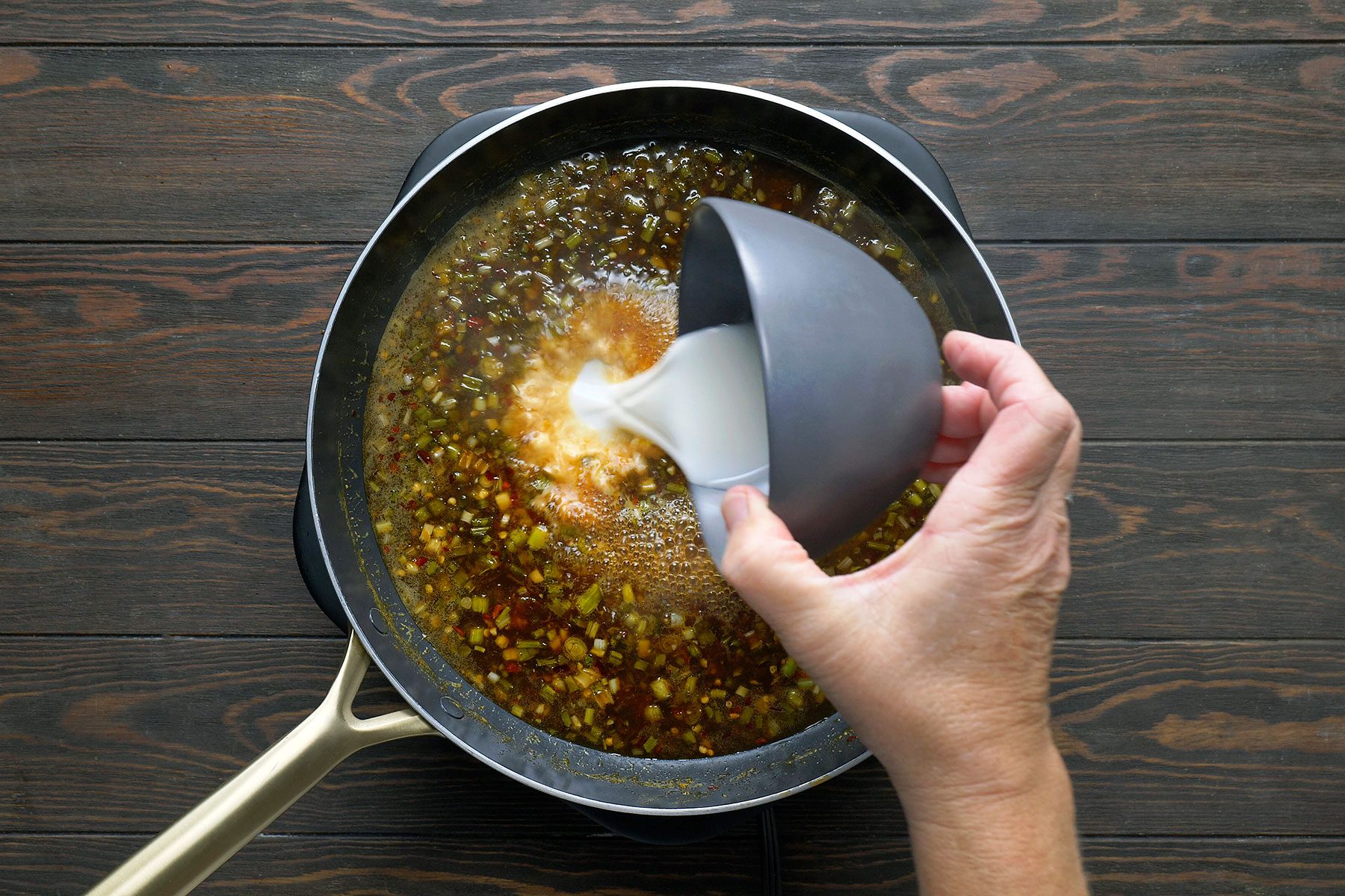 A person pours a creamy liquid from a gray bowl into a frying pan containing a simmering mix of spices and ingredients. The pan rests on a dark wooden surface.