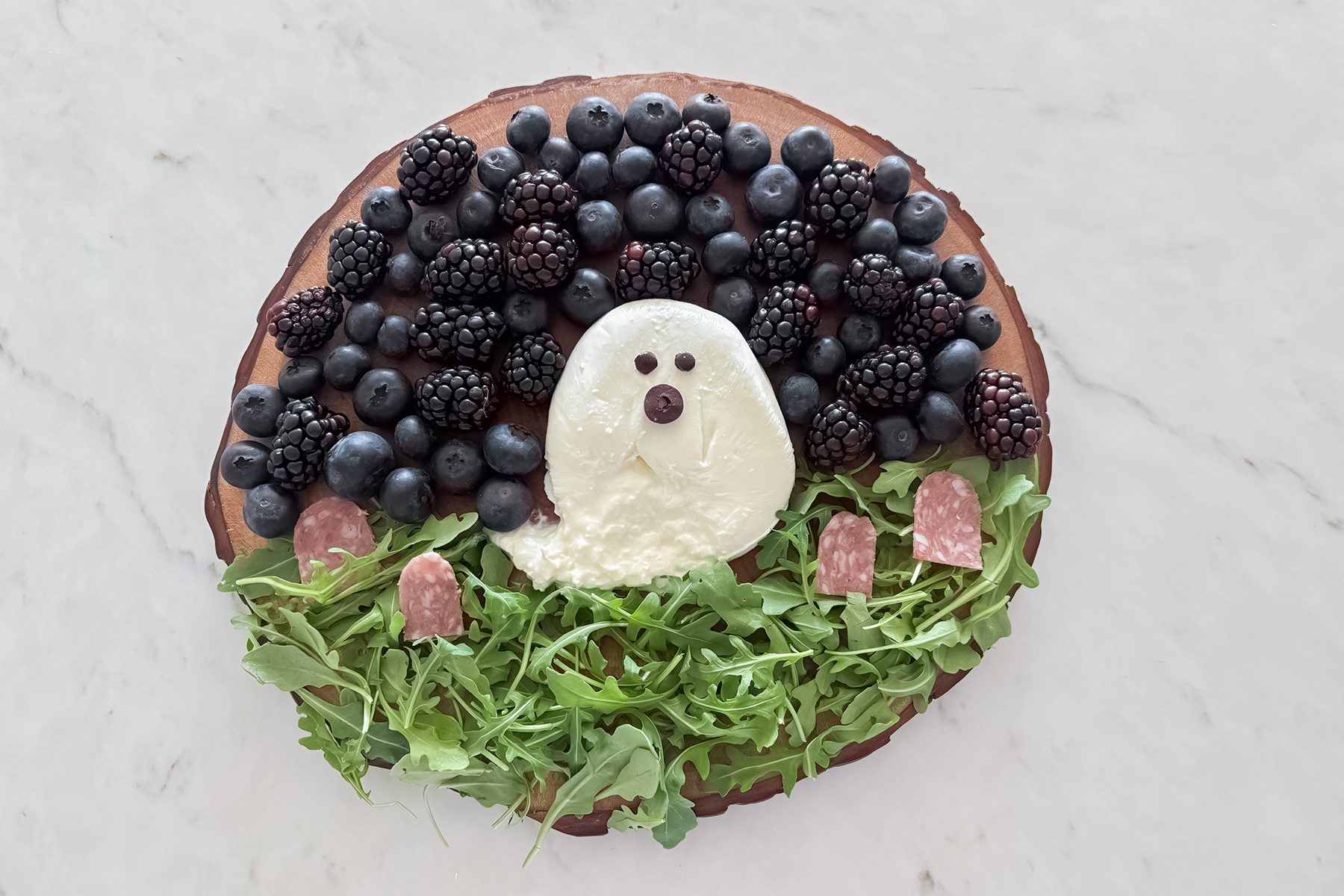 A creative platter shaped like a sheep using blueberries and blackberries as wool, a cheese body with berry eyes and nose, salami slices as feet, and a bed of arugula for grass, on a wooden board.