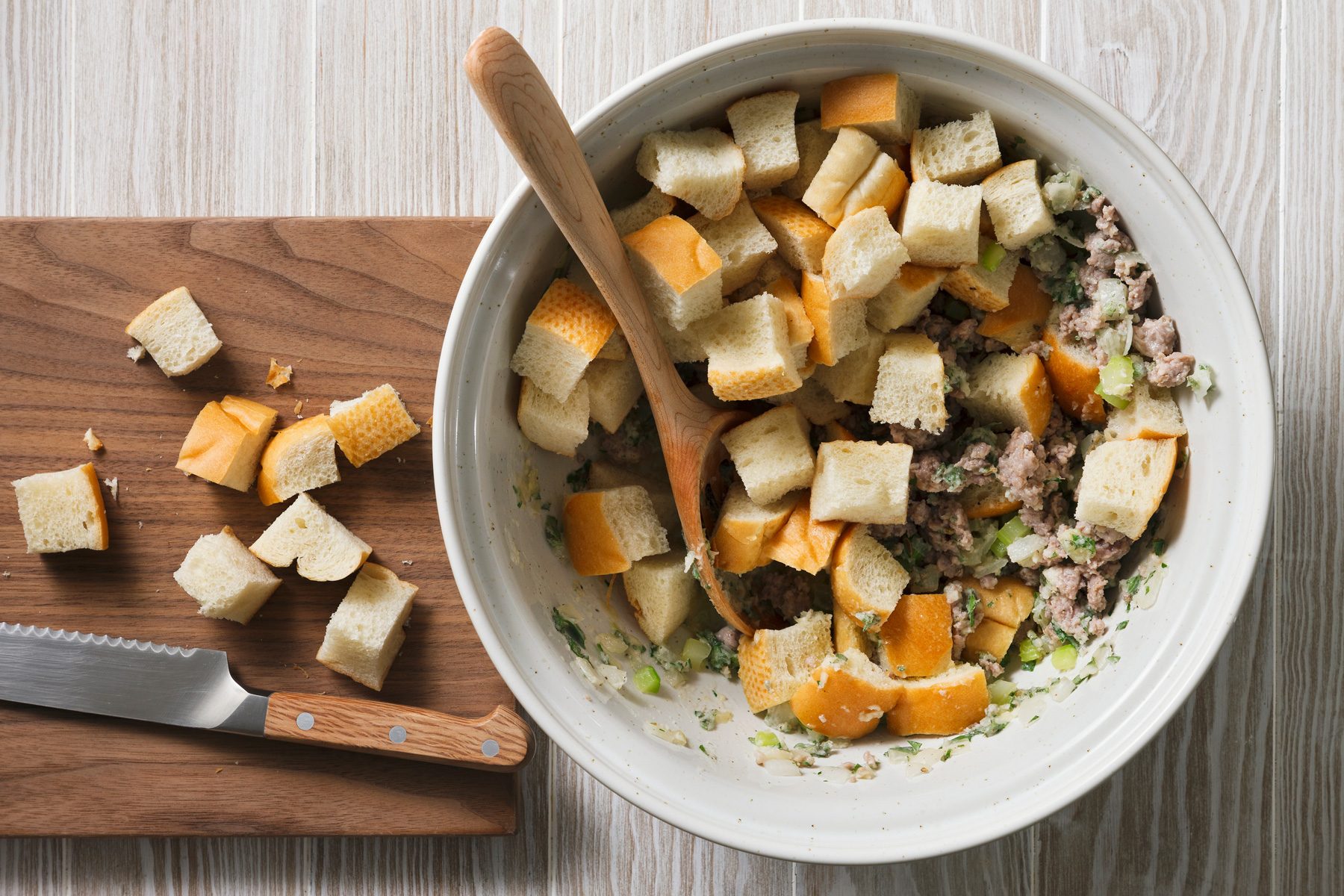 Overhead shot of a bowl filled with cubed bread, ground meat, and chopped celery and onion, There's a wooden spoon partially in the bowl, There are also pieces of bread on a wooden cutting board beside the bowl, A knife is laid on top of the cutting board.