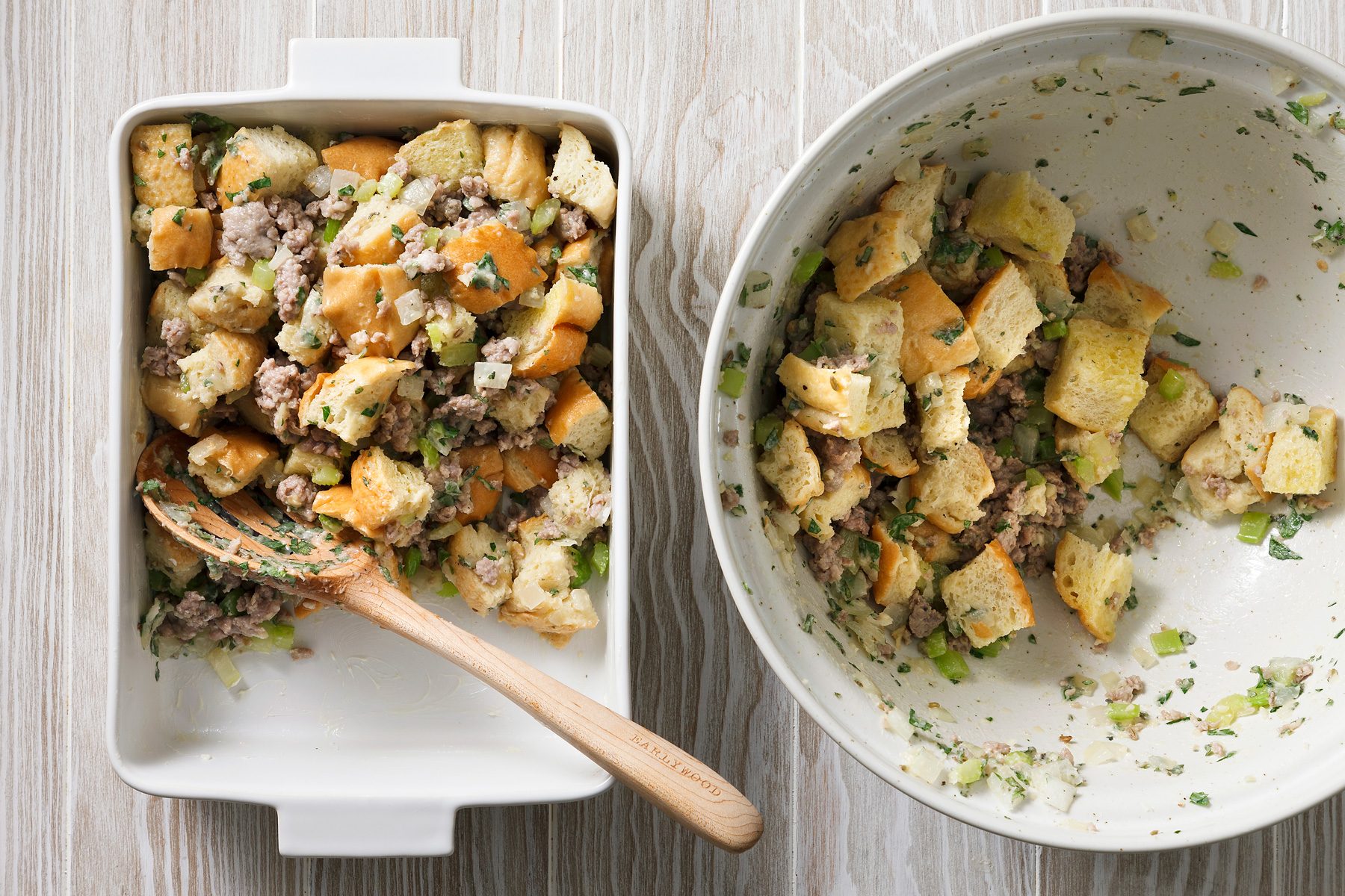 Overhead shot of two white dishes, one with a wooden spoon in it, on a white wooden background, Both dishes contain a mixture of sausage and bread cubes, The dish with the spoon is full, while the other dish is mostly empty, with only a few crumbs of bread and sausage remaining.