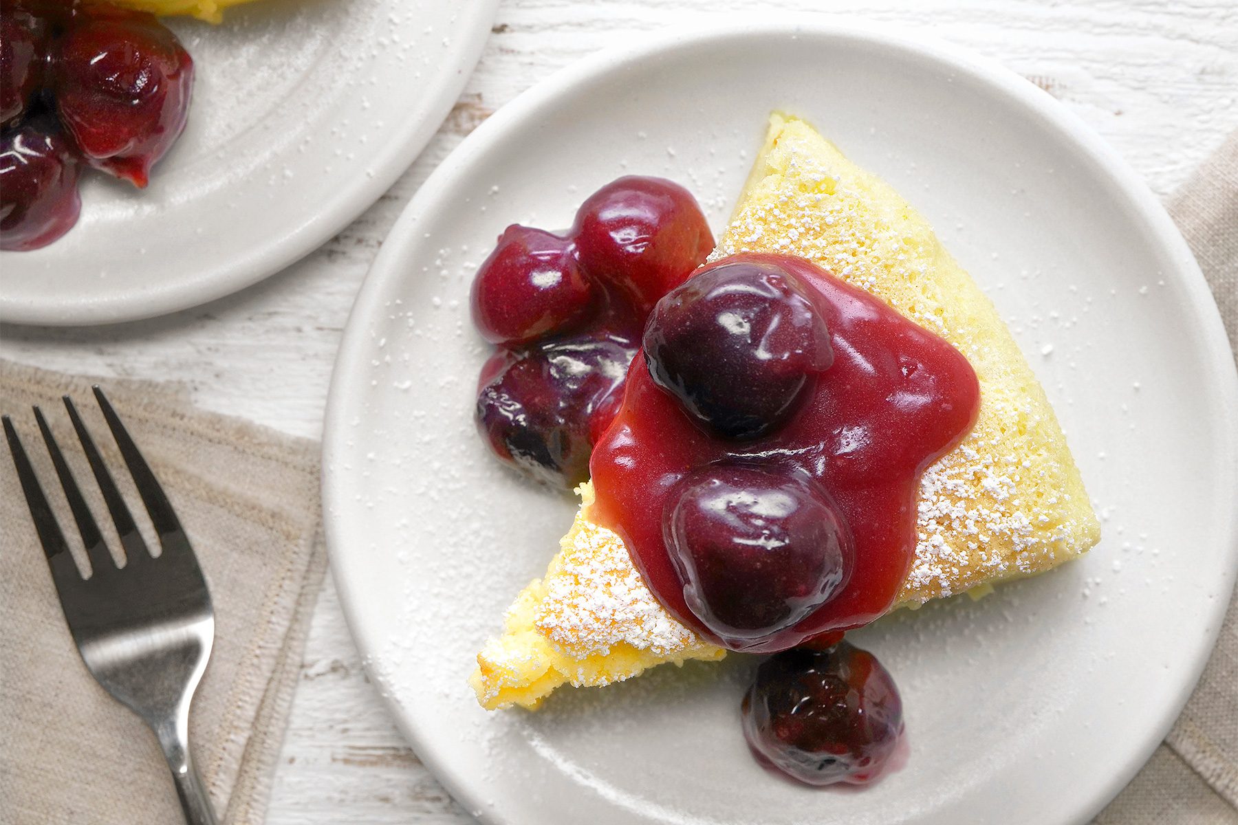 overhead shot of a slice of Japanese Cheesecake is presented on a white plate, topped with a generous dollop of cherry compote, The cherry compote adds a vibrant burst of color and a sweet, tangy flavor, a sliver fork is placed near the plate over kitchen napkin;