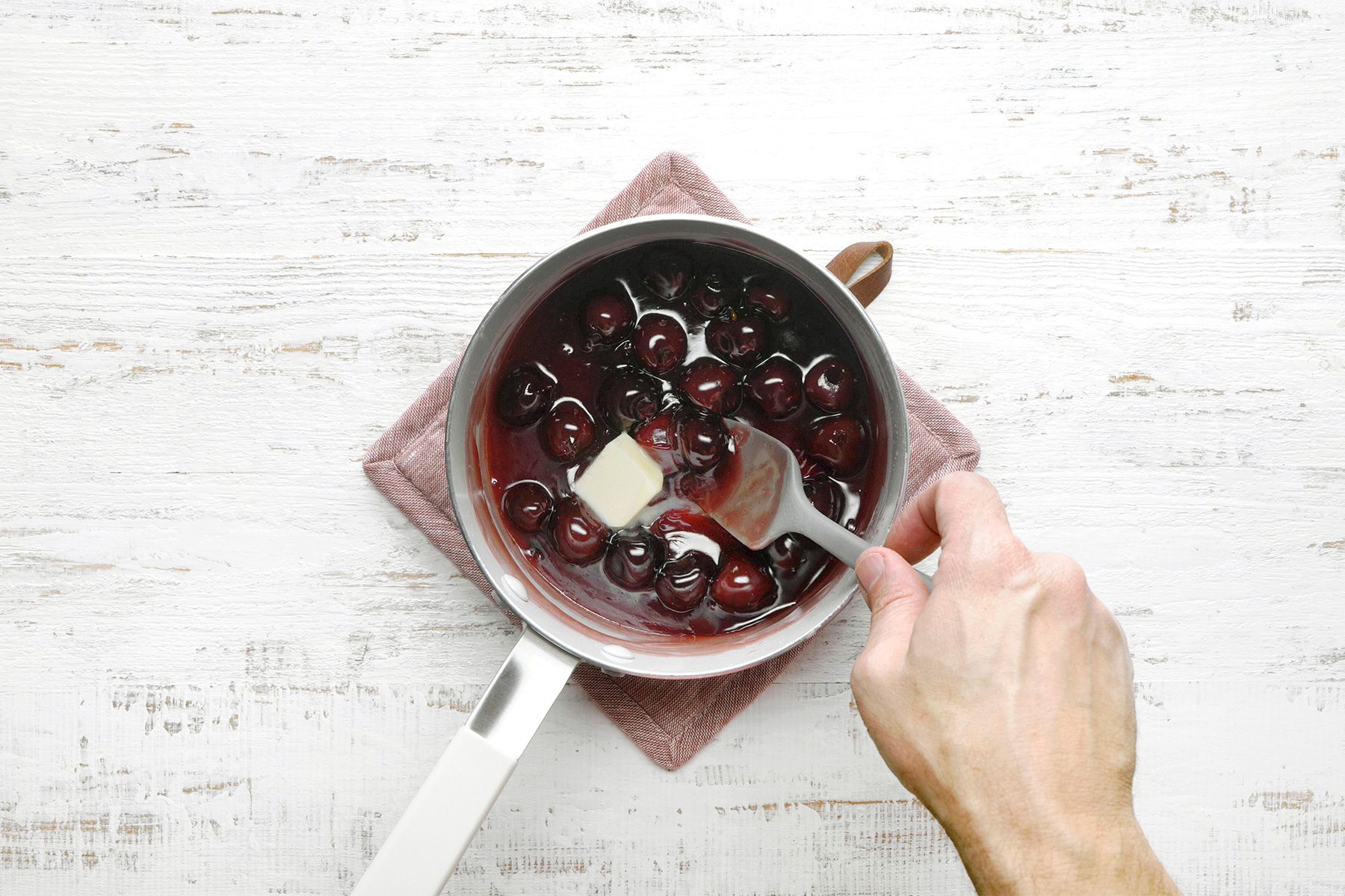 overhead shot of a saucepan contains a mixture of cherries and a reddish-orange liquid, A cube of butter is being added to the mixture and stirred with a spatula;