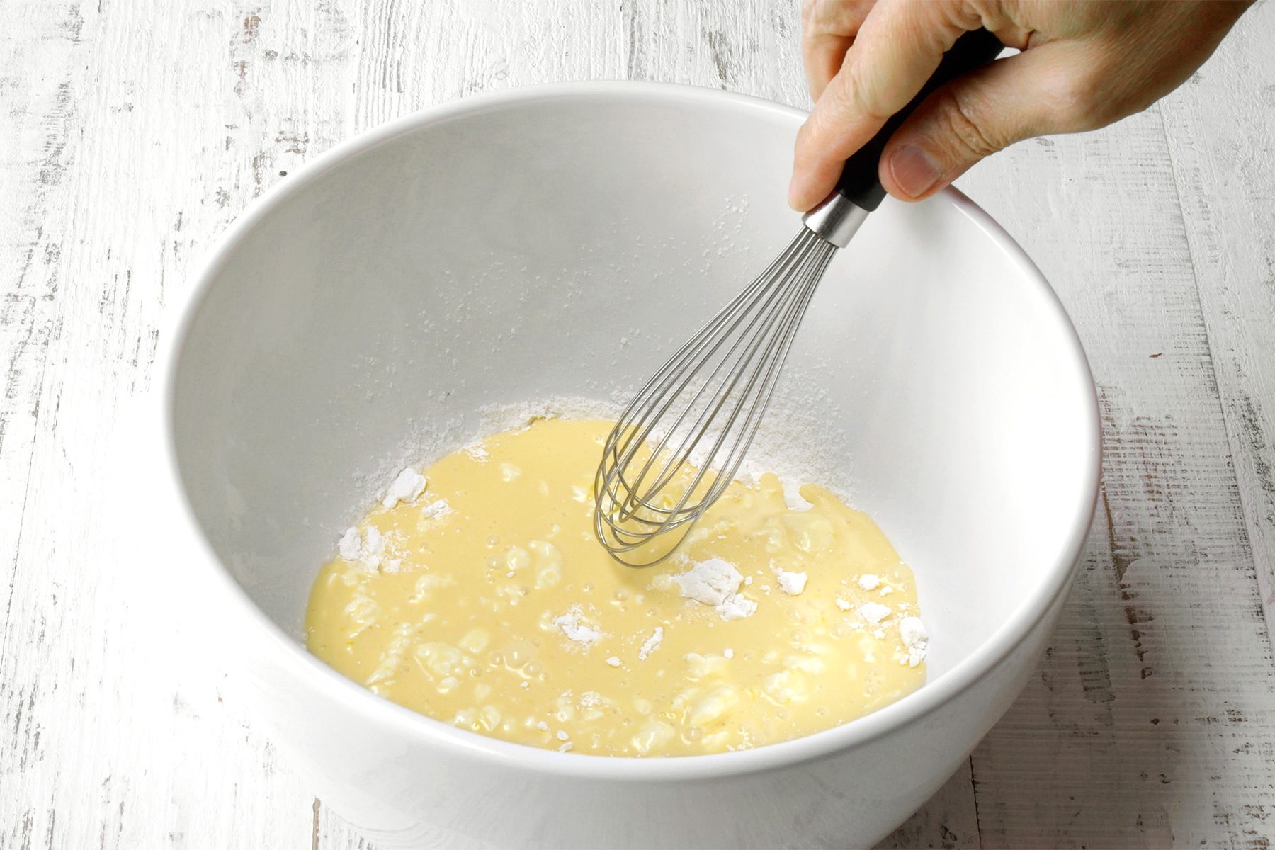 3/4th shot of a hand holding a whisk and whisking a mixture in a white bowl, The mixture appears to be a light yellow color and has a slightly lumpy texture, The bowl is placed on a wooden surface;
