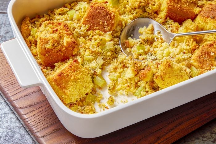 3/4th shot of a white baking dish filled with golden brown cornbread stuffing is sitting on a wooden cutting board, The stuffing is dotted with small pieces of celery and sage, A spoon is laying in the upper left hand corner of the photo, Behind the cutting board, a white plate with a green napkin is in the lower right corner, The background is a dark gray speckled countertop.