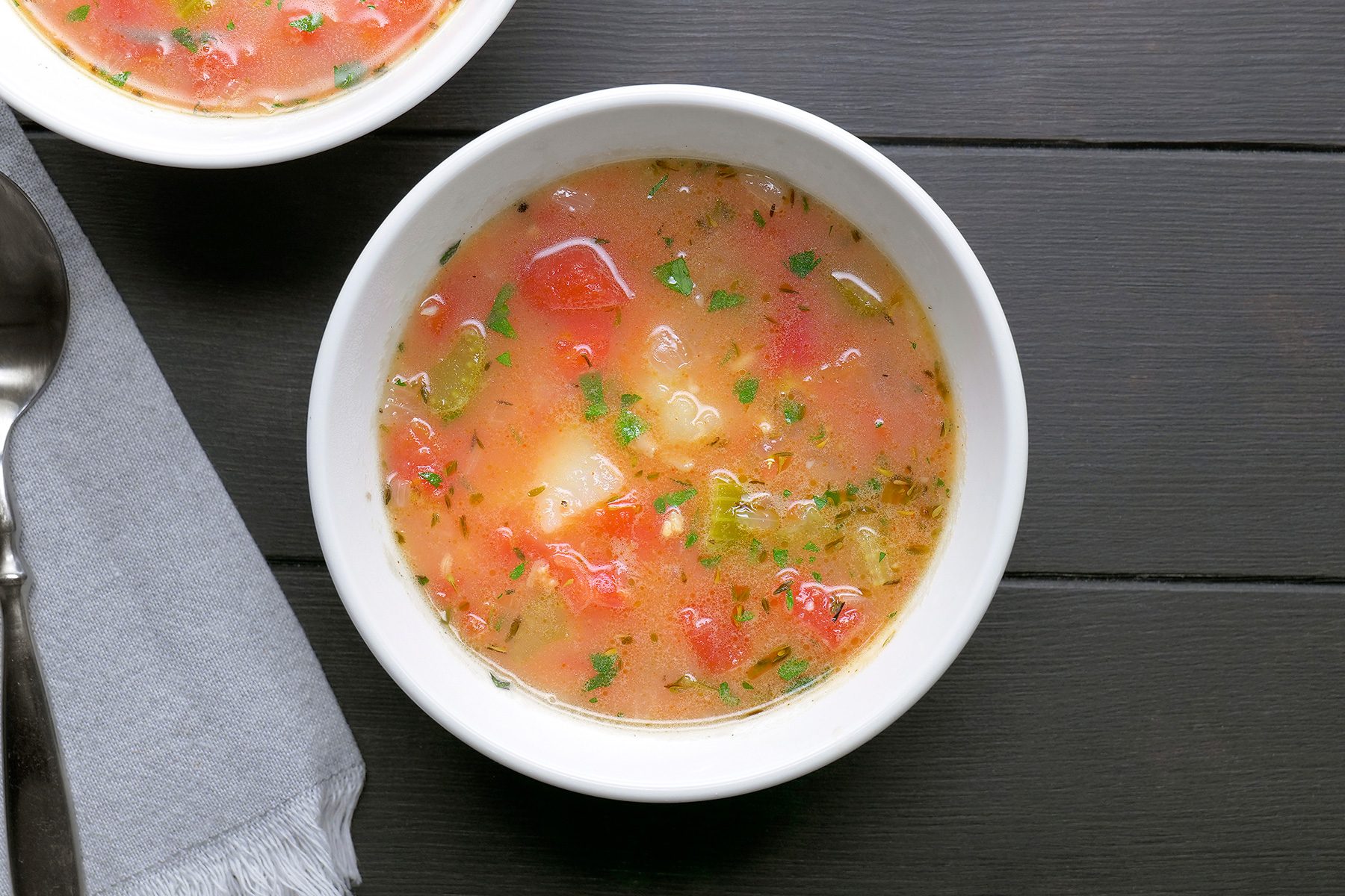 A bowl of colorful vegetable soup with chopped tomatoes, herbs, and green vegetables, placed on a dark wooden table next to a neatly folded gray napkin and a silver spoon.