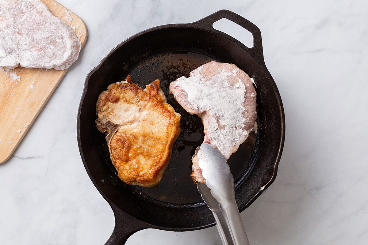 Flour-coated pork chops being fried in butter in a cast iron skillet for step two of Maple-Glazed Pork Chops recipe for Taste of Home