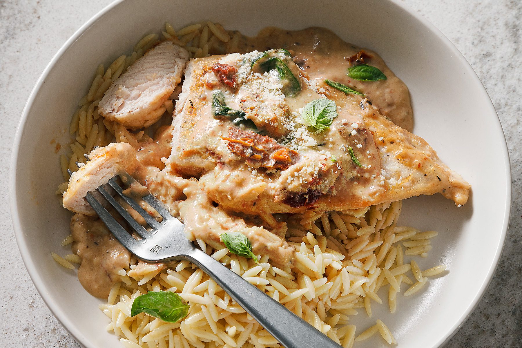 overhead shot of a white bowl filled with a creamy, delicious looking dish, It has orzo pasta, chicken, and a creamy sauce, The sauce has sun-dried tomatoes, basil, and parmesan cheese sprinkled on top, There is a fork on top of the dish, The bowl is on a counter top with a white speckled surface;