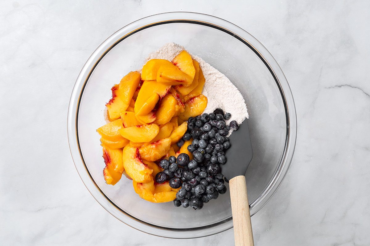 Sugar, flour, cinnamon and allspice in a mixing bowl with added sliced peaches and blueberries for step one of Peach and Blueberry Pie for Taste of Home