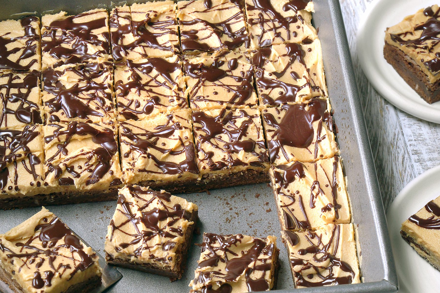 A pan of chocolate-drizzled peanut butter bars cut into squares. One piece is placed on the side, while two white plates are partially visible on the right. The bars have a creamy, peanut butter layer on top.