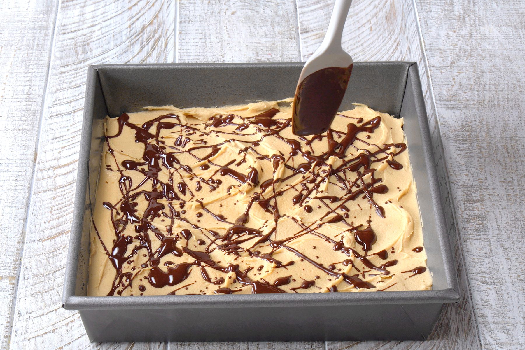A square baking pan filled with light brown batter is being drizzled with melted chocolate. A spatula is spreading the chocolate on the surface. The pan is placed on a light wooden surface.