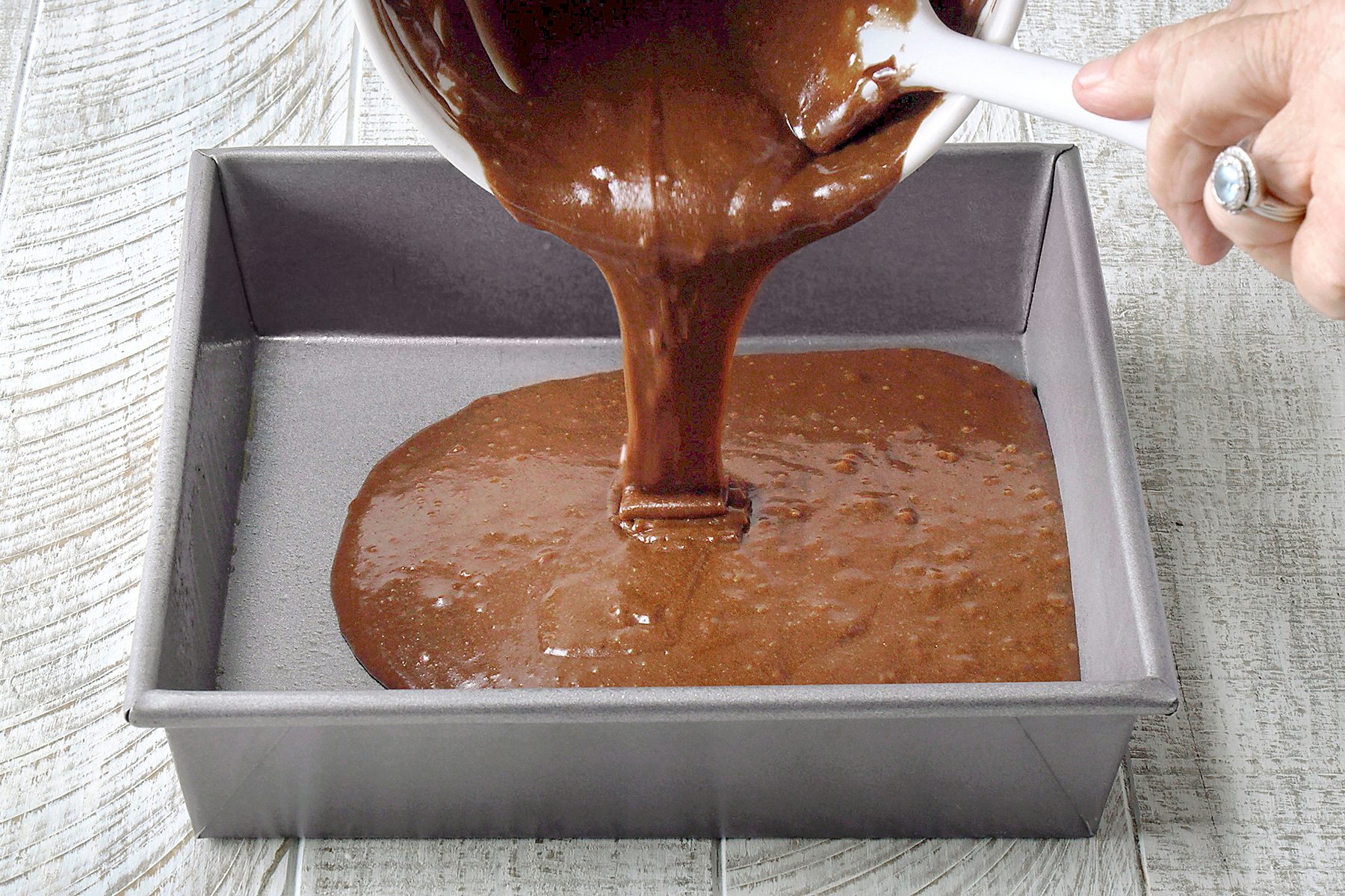 A person pours chocolate cake batter from a white bowl into a greased square baking pan on a wooden surface.