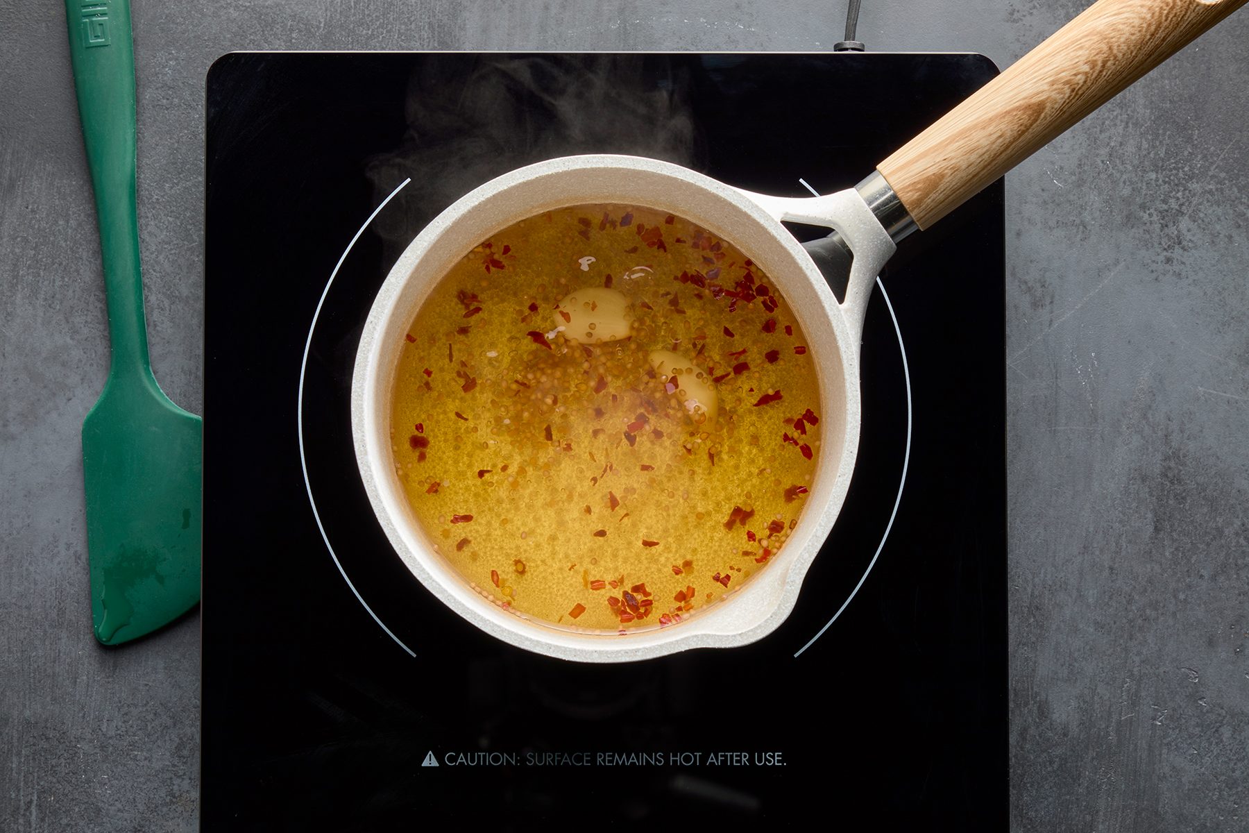A pot with broth containing spices and seasoning simmers on an induction cooktop. A wooden-handled spatula is placed beside the cooktop on a dark gray surface. Steam is visible rising from the pot.