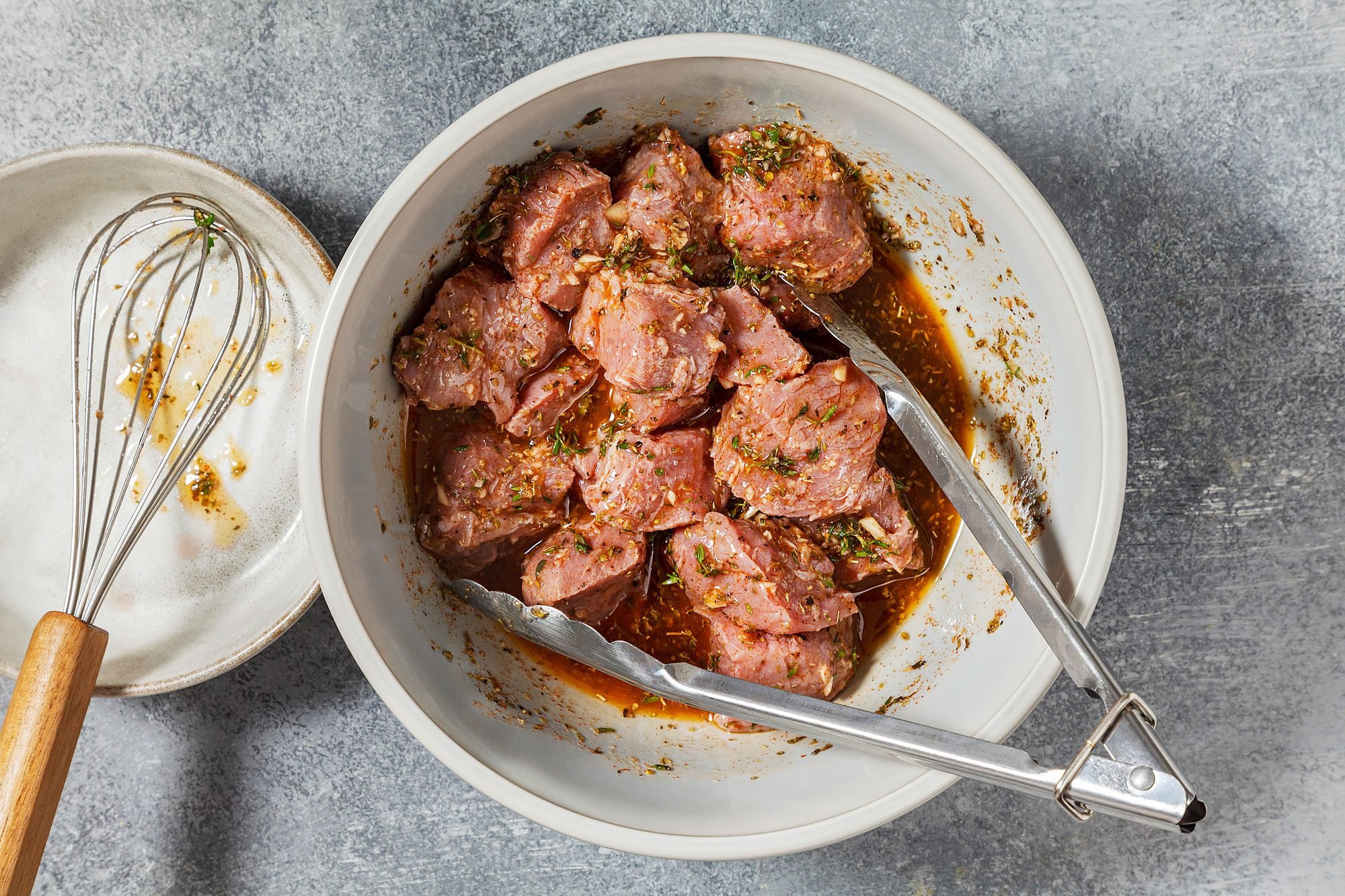 overhead shot of a white bowl filled with marinated meat, The meat is cut into small cubes and is coated in a reddish-brown marinade, The marinade appears to be a mixture of spices and liquids, A pair of tongs is resting in the bowl, and a whisk is placed on the side;