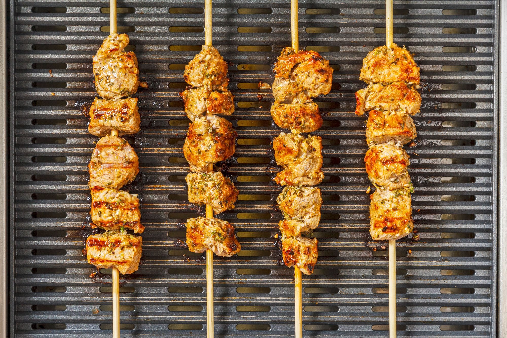 overhead shot of four skewers of grilled meat on a grill, The meat is cooked to a golden brown color and is slightly charred, The skewers are arranged in a parallel pattern on the grill;