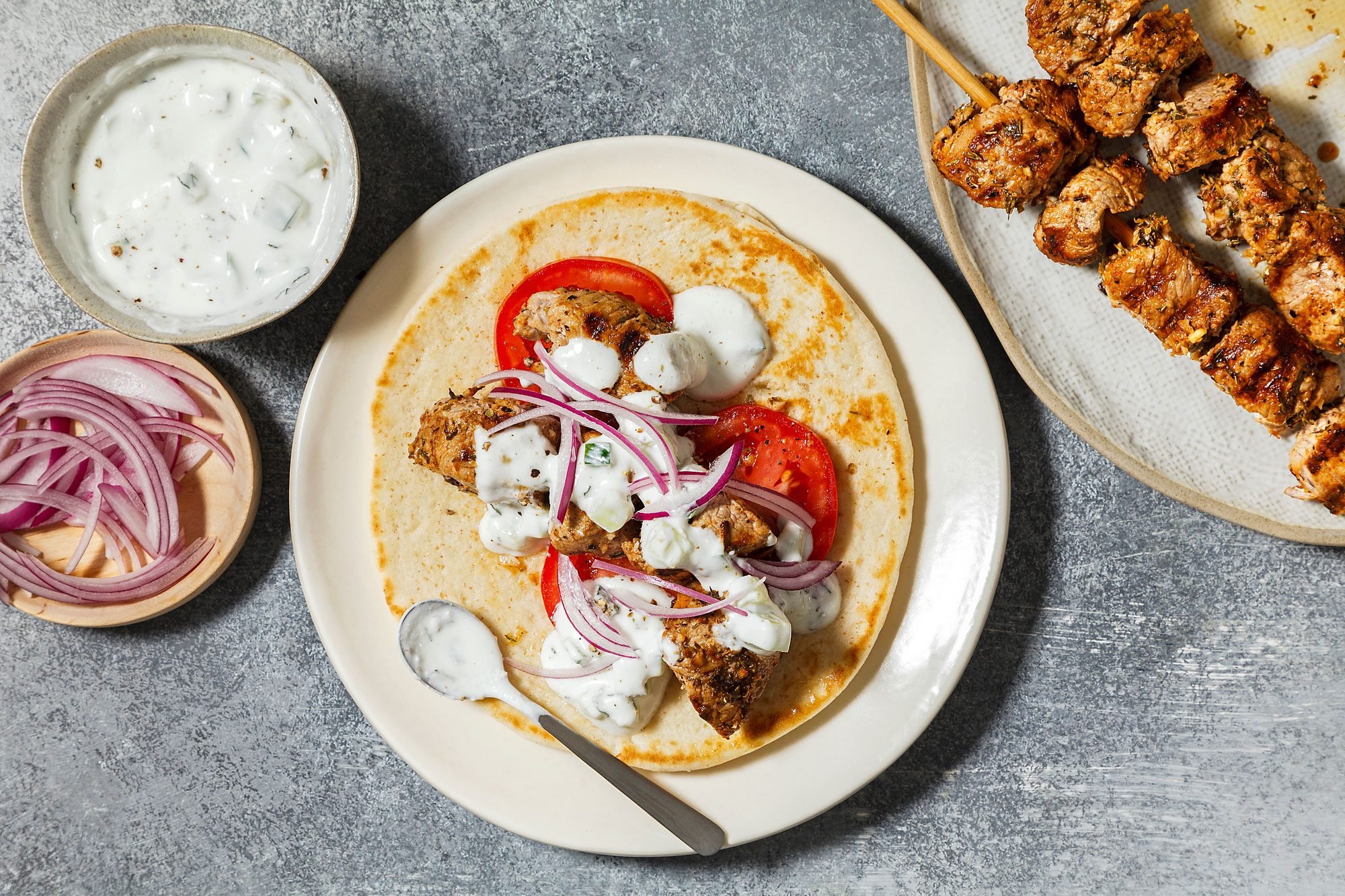 overhead shot of a plate of Greek gyros, A pita bread wrap is filled with grilled chicken skewers, sliced tomatoes, red onion, and a generous dollop of tzatziki sauce, The chicken is cooked to a golden brown color and is seasoned with herbs and spices, The tzatziki sauce is creamy and white, and the vegetables are fresh and colorful;