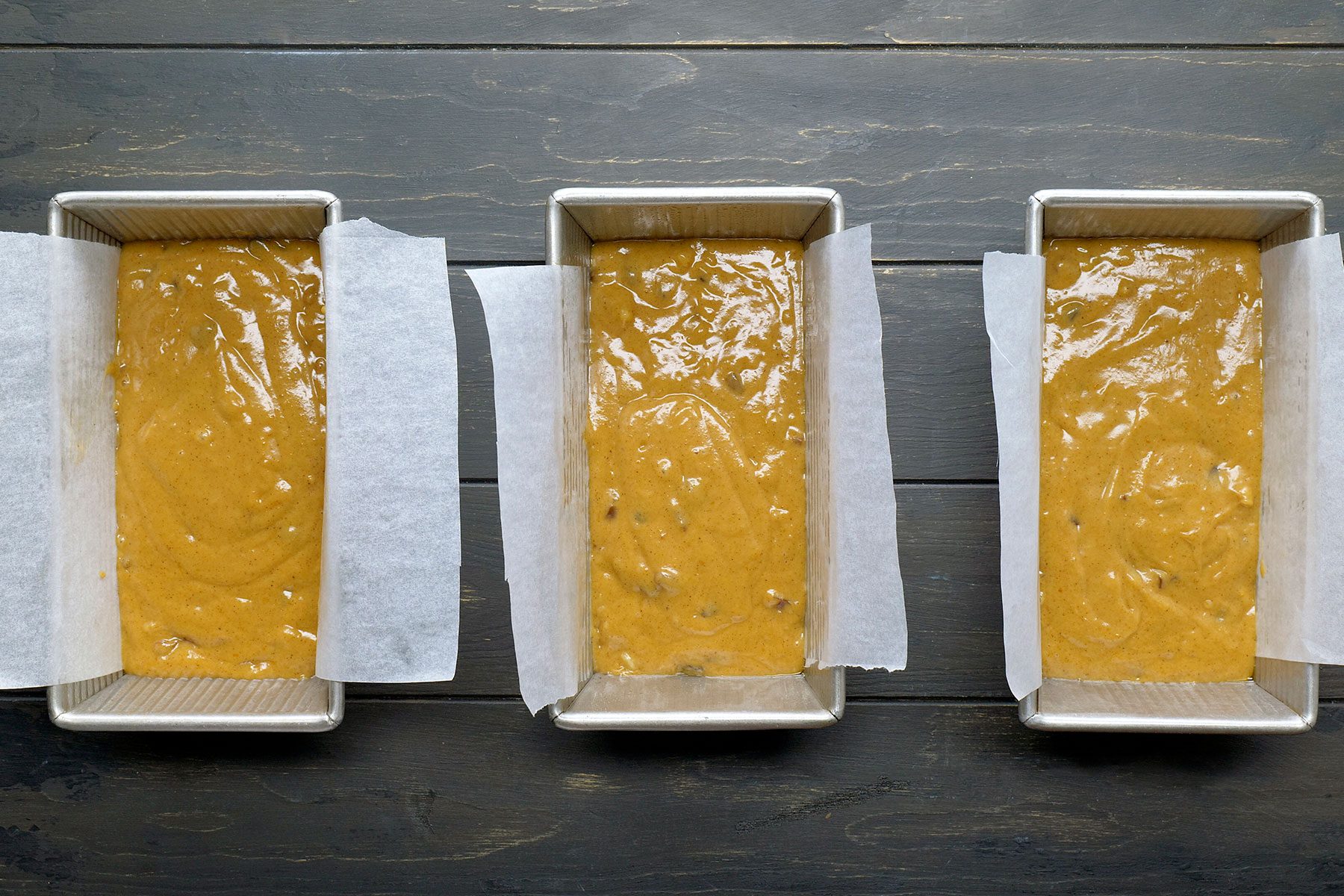Three rectangular baking pans lined with parchment paper, filled with unbaked batter, sit on a dark wooden surface. The batter is golden brown, suggesting a sweet loaf or bread in preparation.