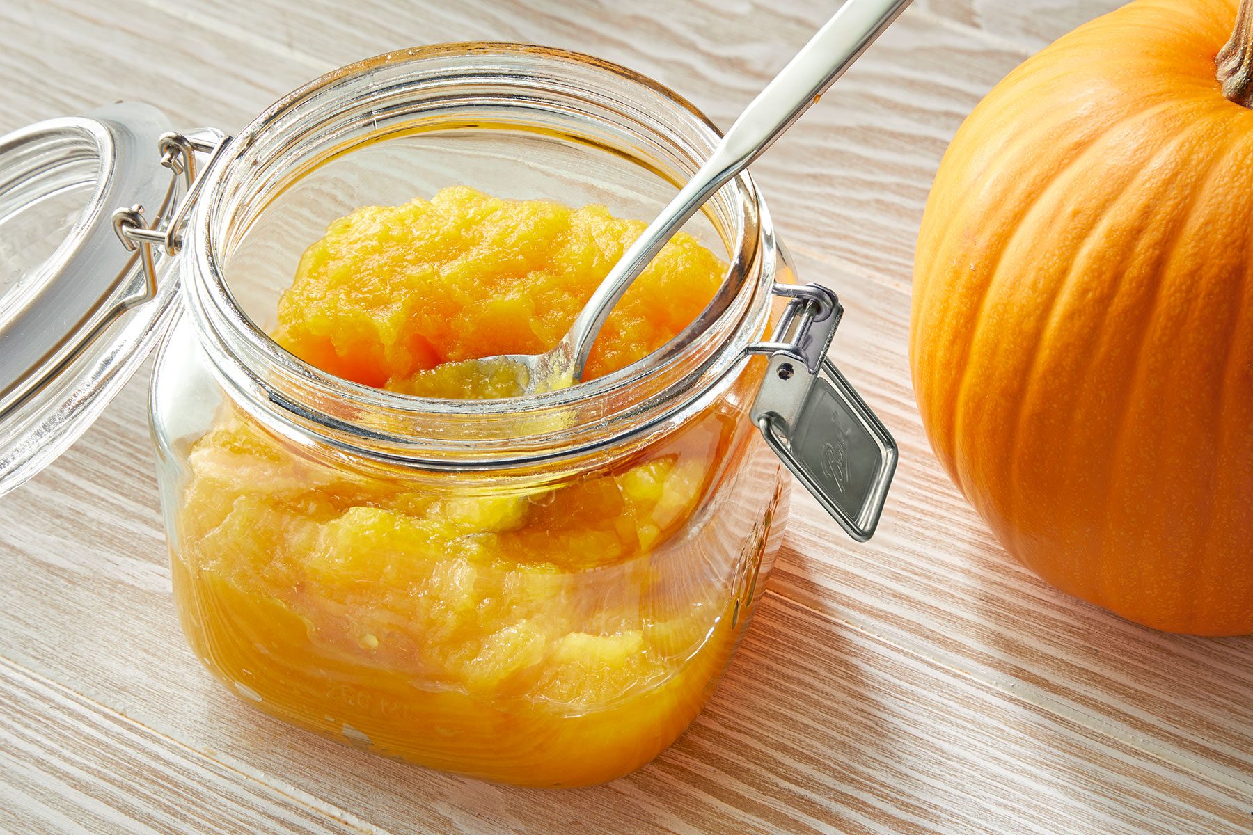 Glass jar of pumpkin puree with a spoon inside, next to a whole pumpkin on a wooden surface.