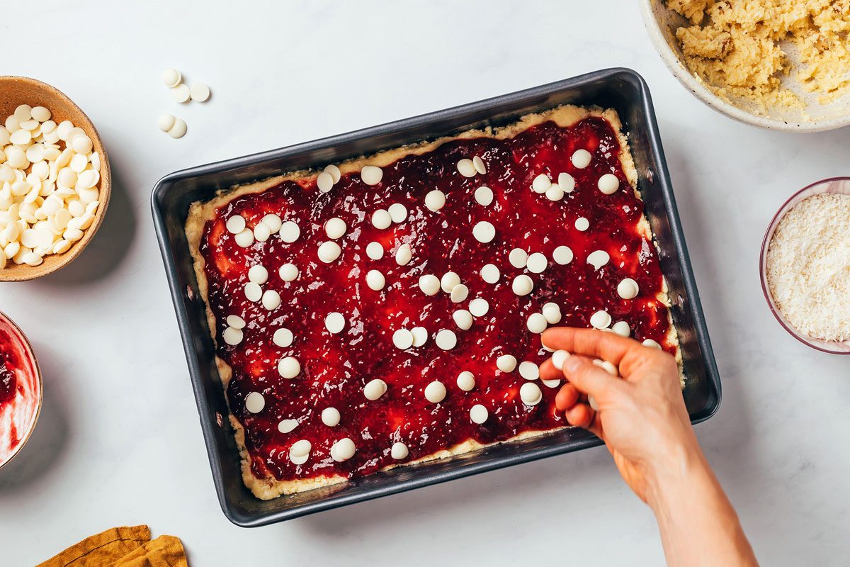 Raspberry bars with coconut being assembled in a baking pan, white baking chips being sprinkled by hand on top of the raspberry preserve layer recipe by Taste of Home