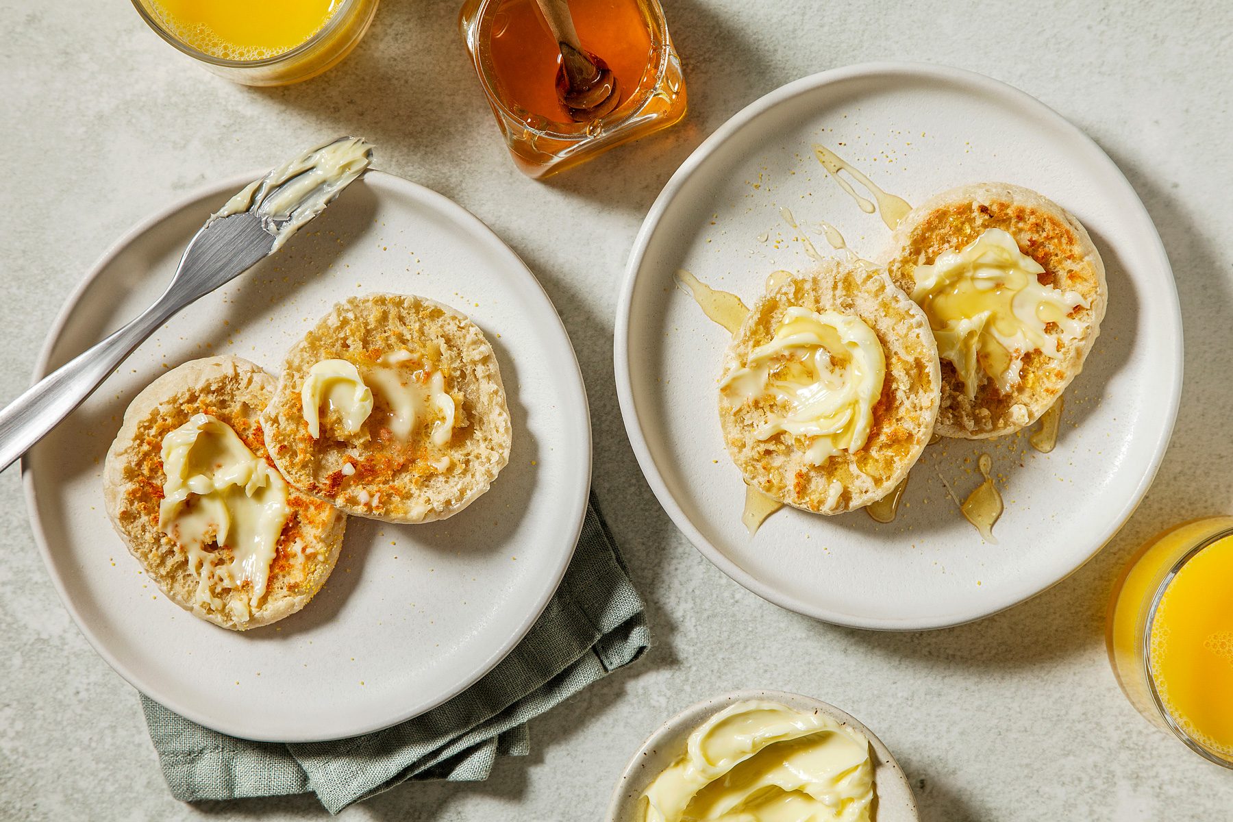 overhead shot of two plates of English muffins, The muffins are split in half and topped with a generous amount of butter and a drizzle of honey, A glass of orange juice is placed on the side of each plate;