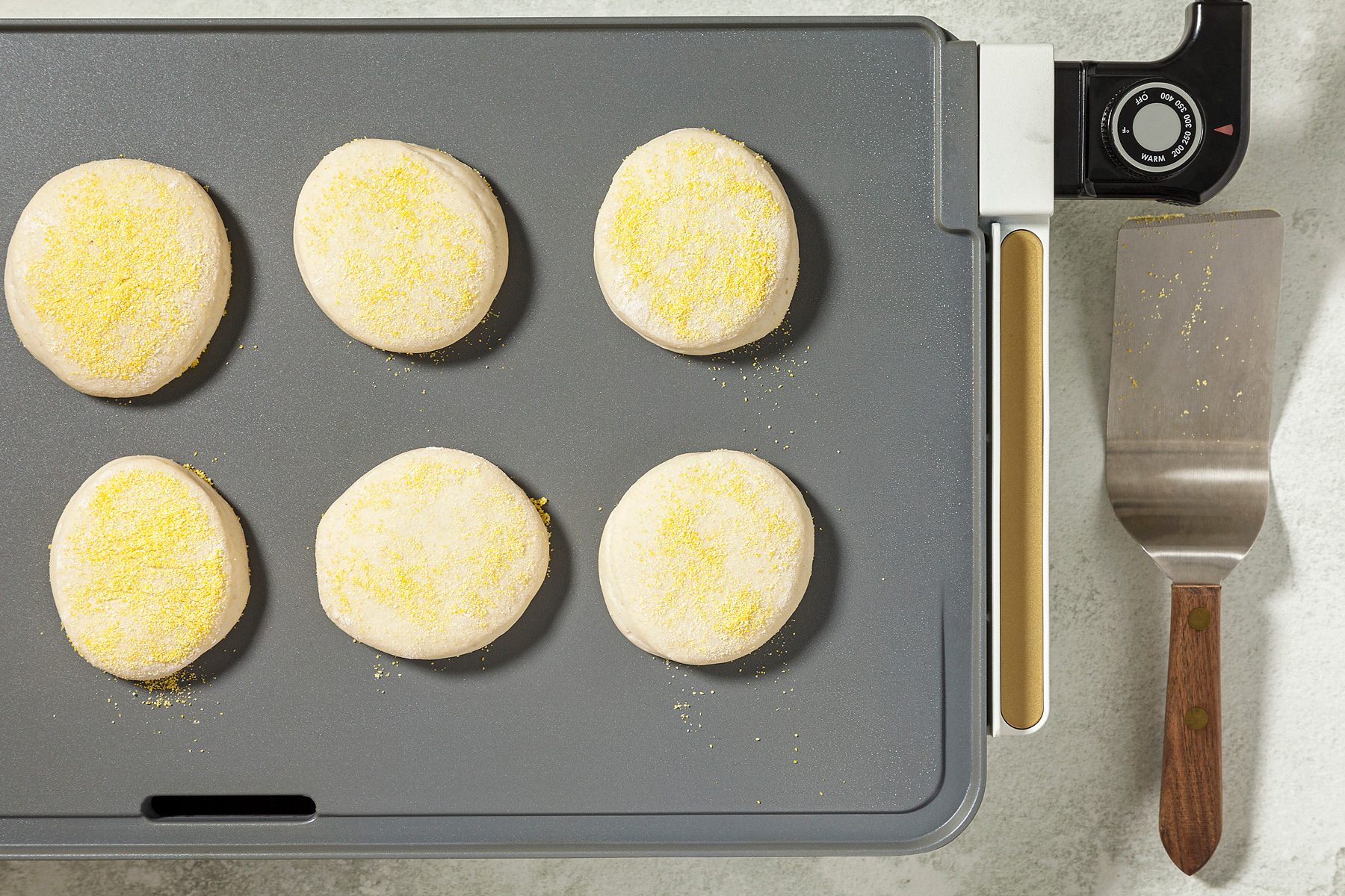 overhead shot of a griddle with six round dough circles arranged in a grid pattern, The dough circles are coated in cornmeal, A spatula is placed on the side of the griddle;
