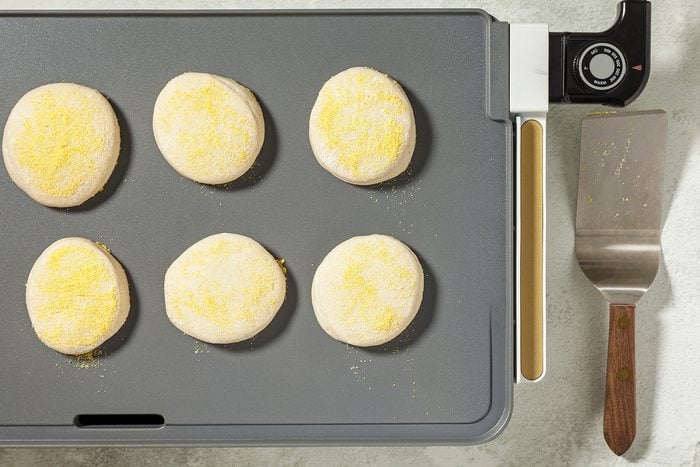 overhead shot of a griddle with six round dough circles arranged in a grid pattern, The dough circles are coated in cornmeal, A spatula is placed on the side of the griddle;