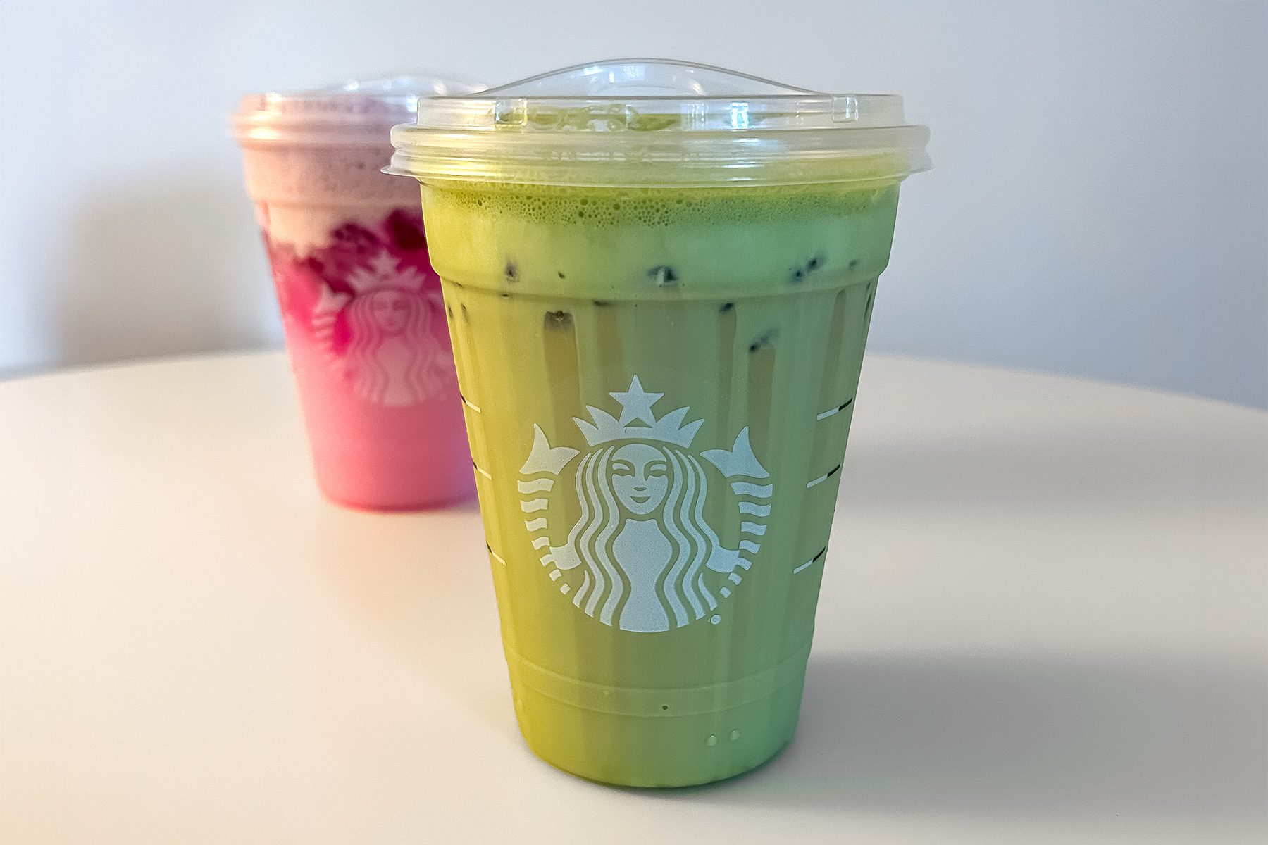 A plastic cup of iced green matcha latte is in the foreground with a clear lid. In the background, there is another pink drink with ice and a similar lid. Both cups have a logo on them, and they sit on a white table.