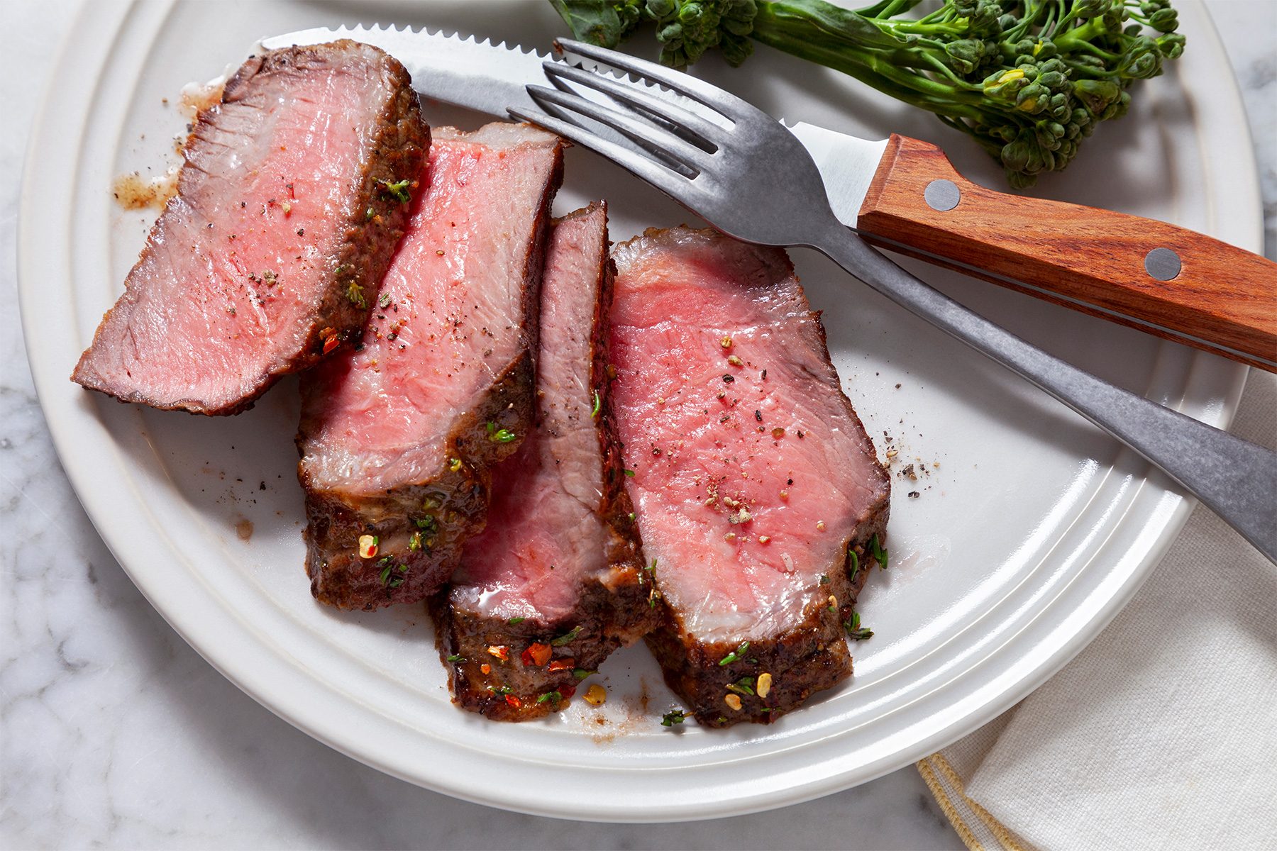 overhead shot of a plate of sliced steak, accompanied by a side of steamed broccoli, The steak is a deep red color and has a slightly charred exterior, It is seasoned with a blend of herbs and spices, The broccoli is a vibrant green and is cooked until tender-crisp, A fork and knife are placed next to the steak;
