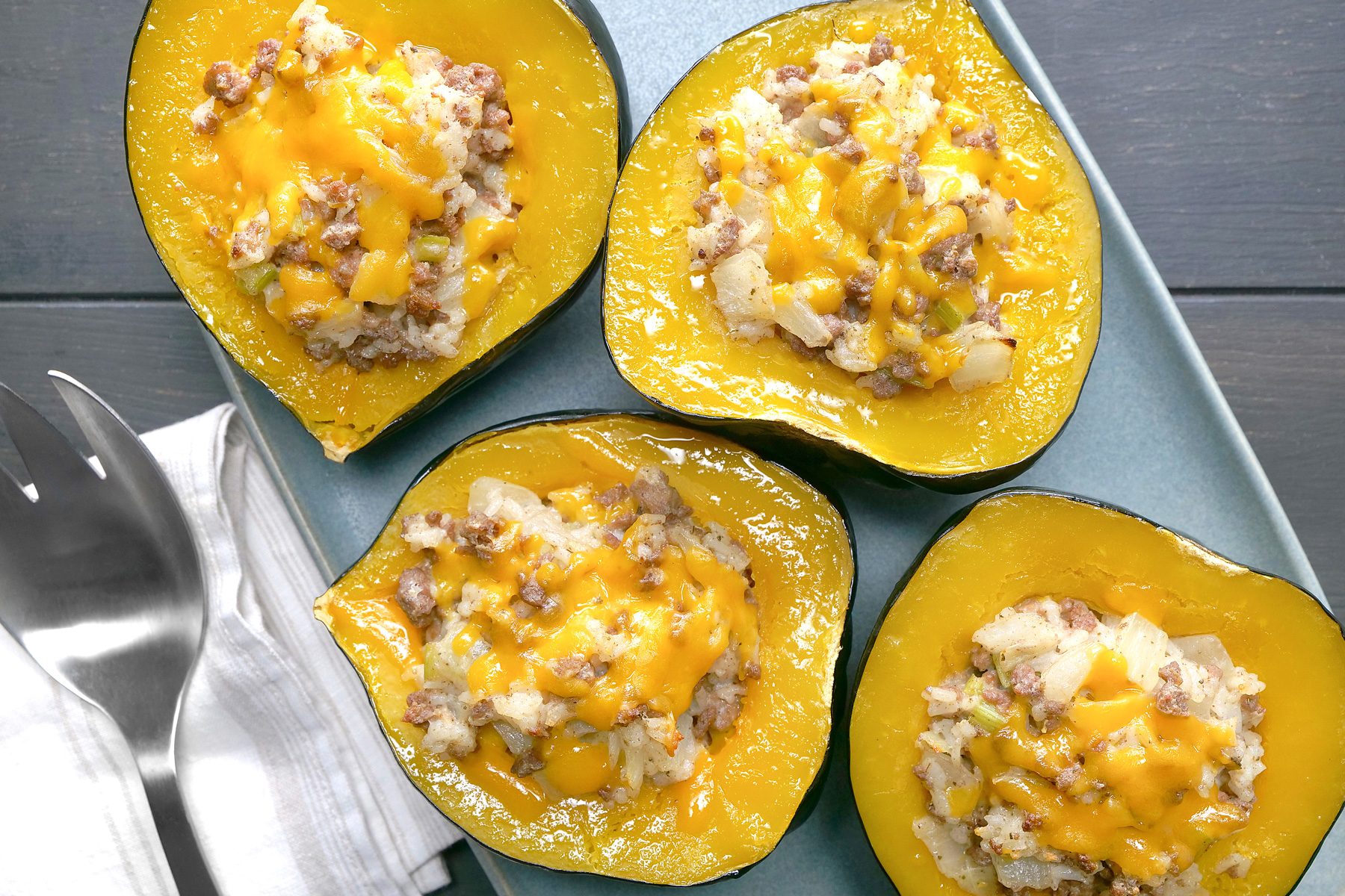 overhead shot of Four acorn squash halves are filled with a mixture of ground meat, rice, and cheese, The meat mixture is a golden brown color and is topped with melted cheese, The acorn squash shells are a vibrant orange color and are still attached to the stems, The squash halves are arranged on a blue platter, and two forks are placed next to the platter;