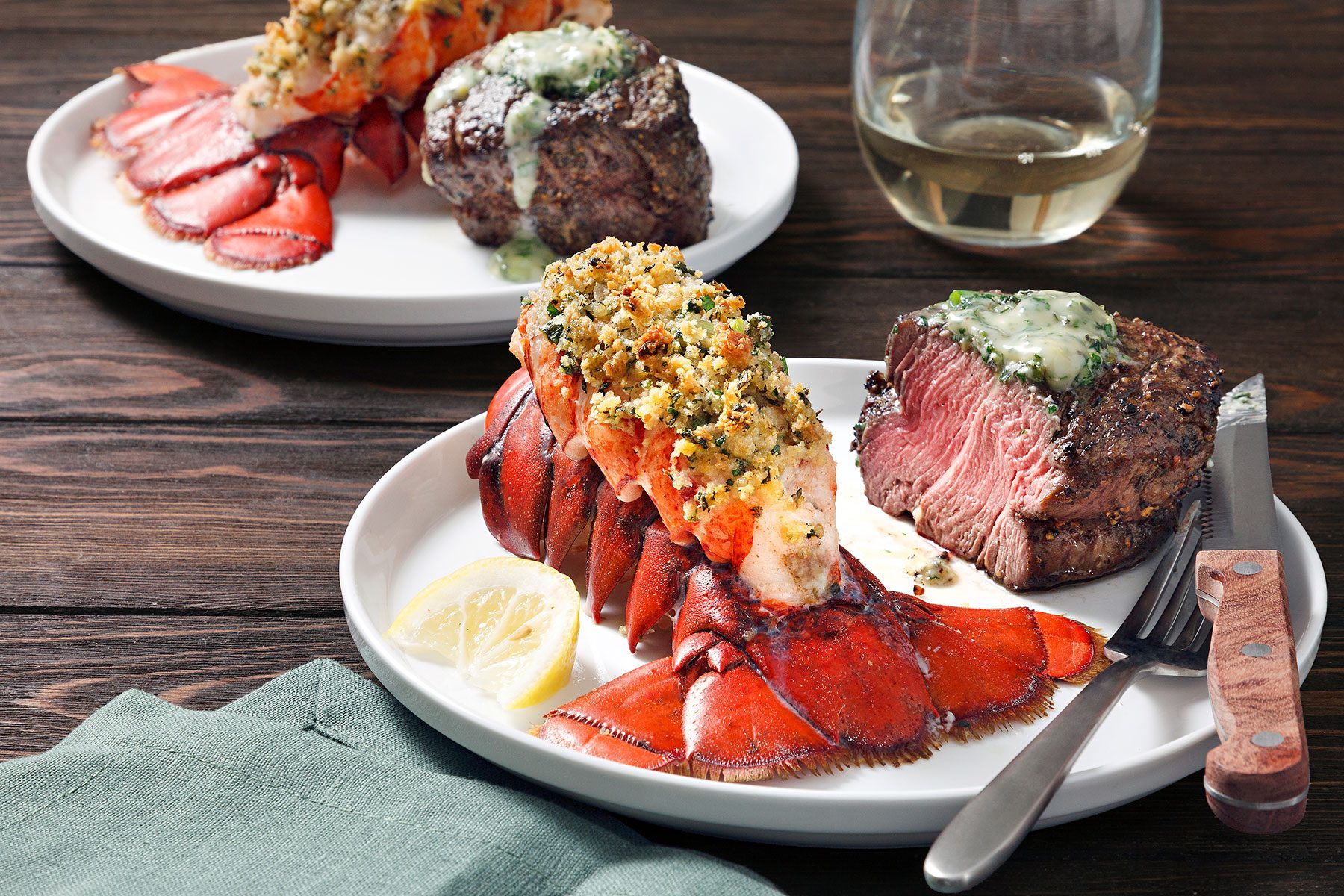 Plated meal with lobster tail topped with herbs and filet mignon, accompanied by a lemon slice, with a second similar plate and a glass of white wine in the background. A fork and knife are set beside a green napkin on a wooden table.
