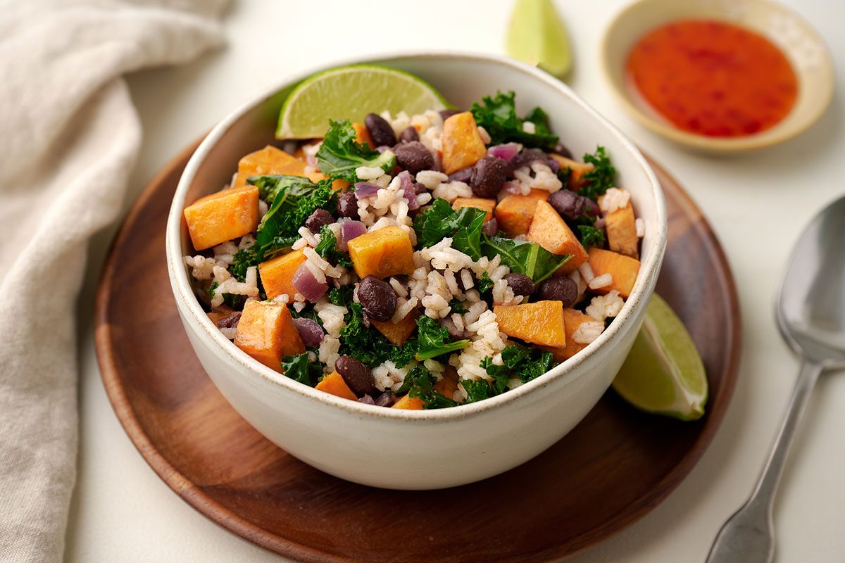 Finished Sweet Potato Bowl with sautéed sweet potatoes, kale, black beans, red onion, and rice, garnished with lime wedges and sweet chili sauce