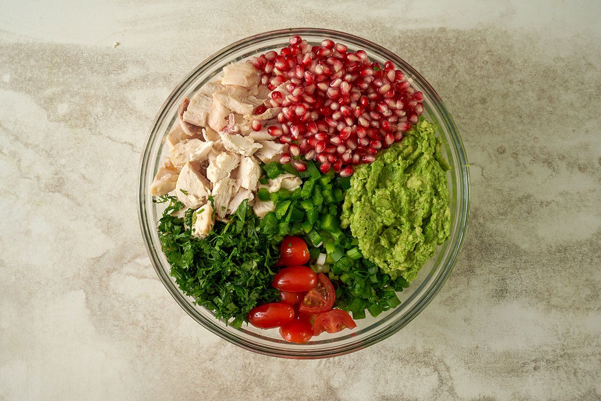 Overhead view of rotisserie chicken, mashed avocados, pomegranate seeds, green onions, cherry tomatoes, jalapeno pepper, and cilantro leaves being combined in a large bowl for the Taste of Home Avocado Chicken Salad recipe.