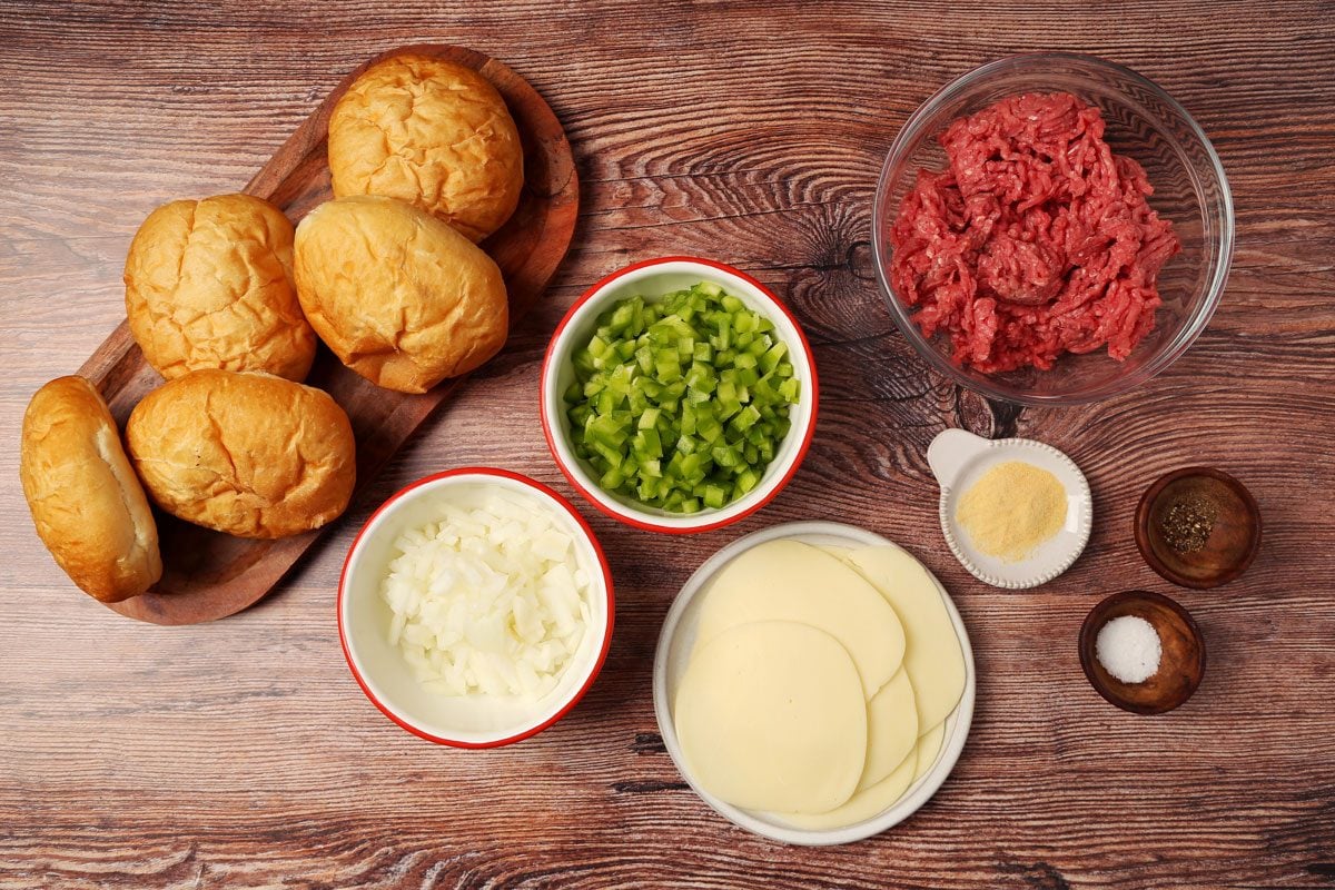 Ingredients for Taste of Home's Philly Cheesesteak Sloppy Joes laid out in small bowls on a brown wooden surface.