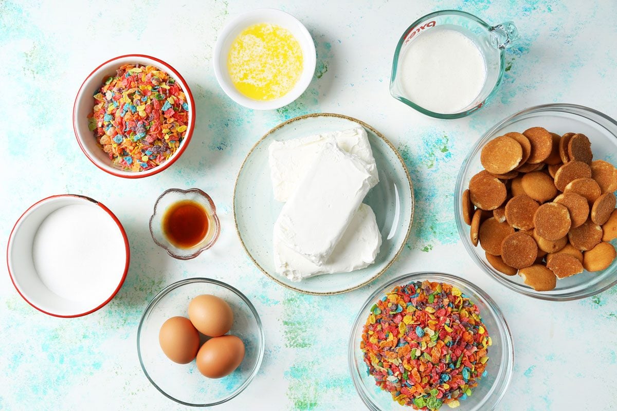 Ingredients for Taste of Home's Fruity Pebbles Cheesecake laid out in small bowls on a blue and green surface.
