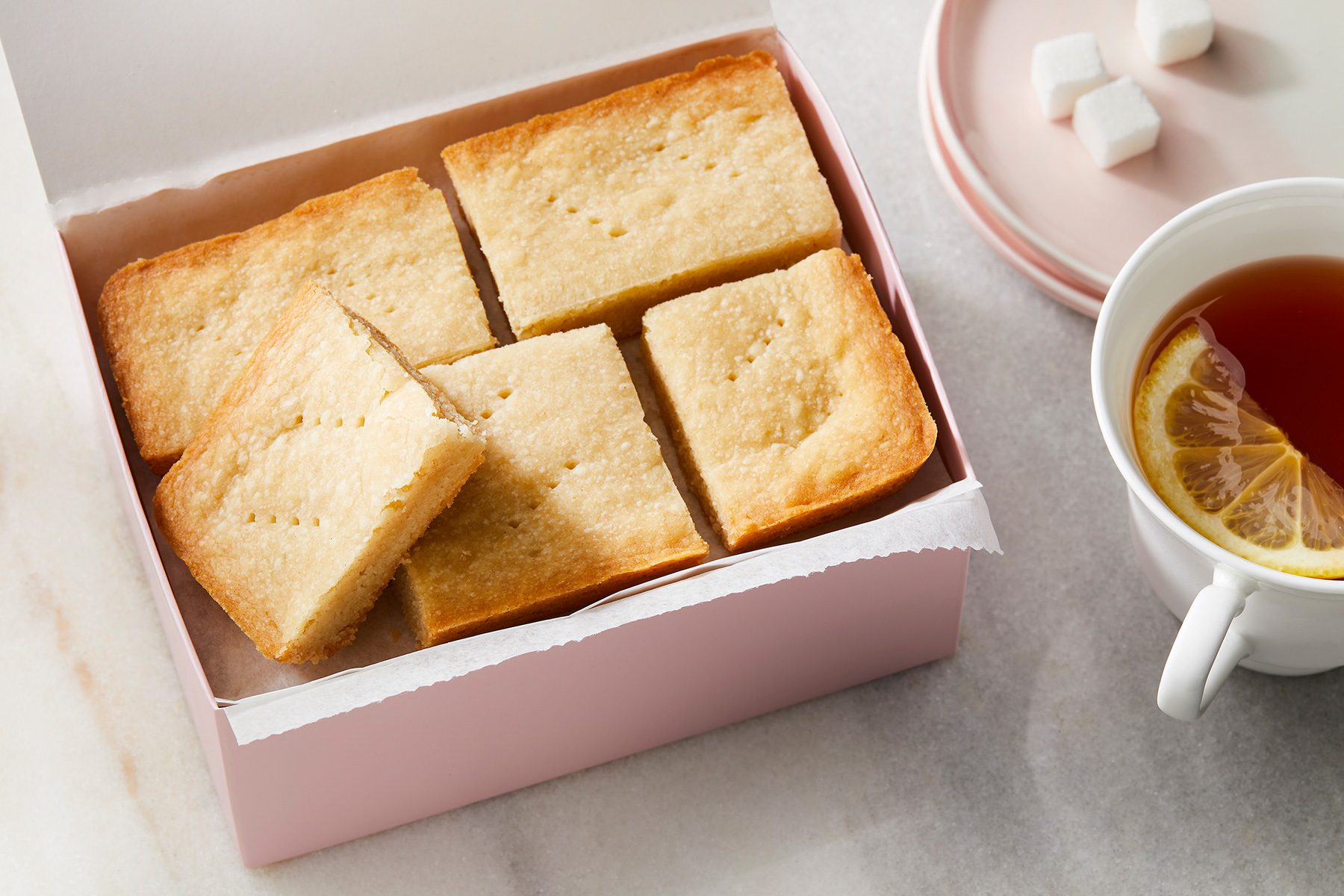 Horizontal AP; Pull shot of a pink gift box filled with Ted Lasso shortbread cookies is showcased, The cookies are square-shaped and have a golden brown color, A cup of tea with a lemon wedge is placed beside the box, along with a small plate of sugar cubes;