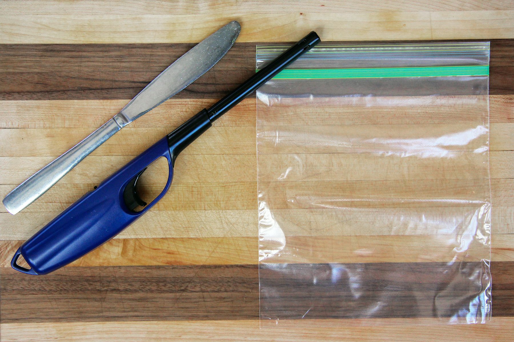 A butter knife, a long lighter, and a clear resealable plastic bag are placed on a wooden cutting board with alternating light and dark wood stripes.